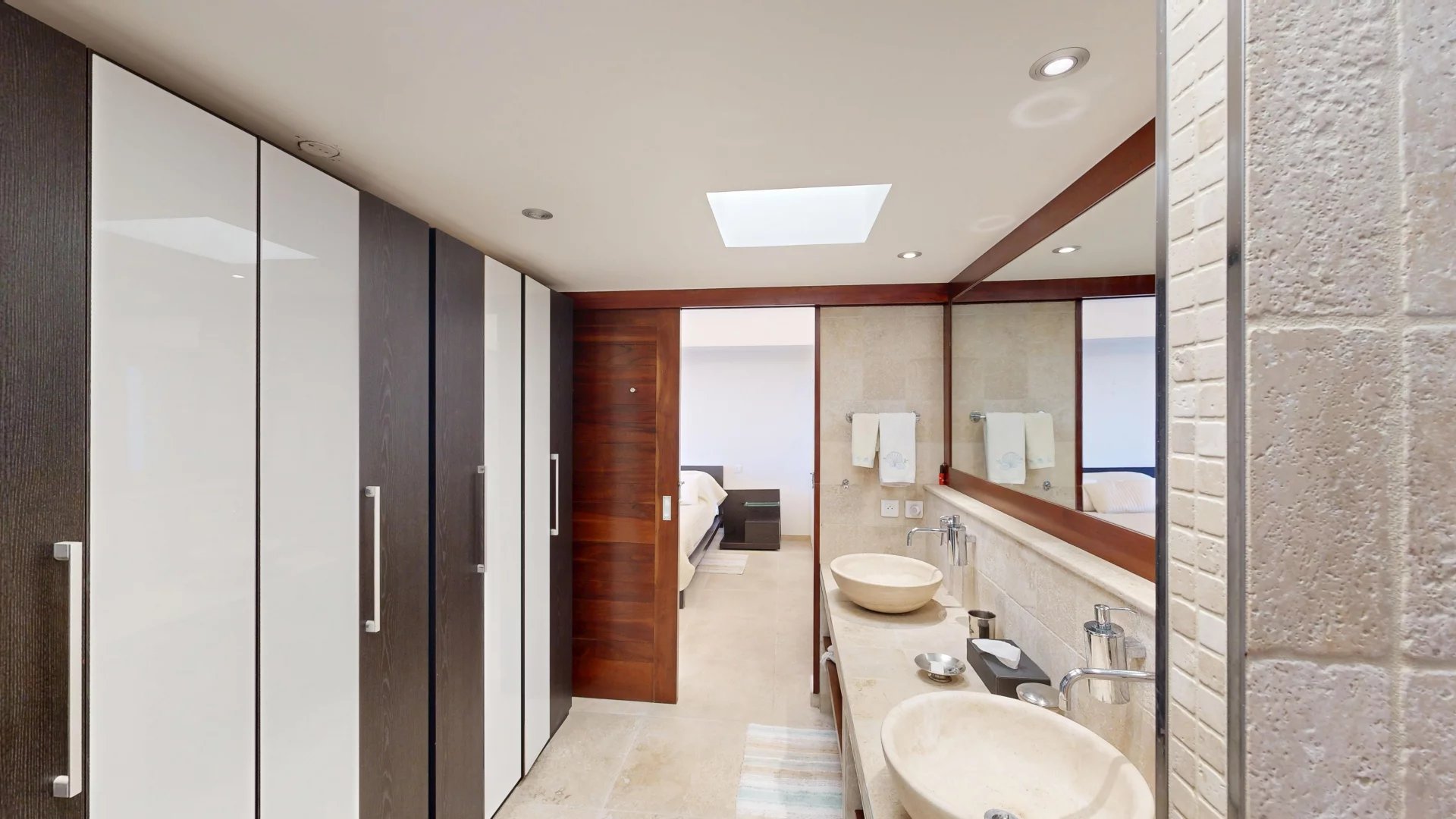 Modern bathroom with a long vanity and two vessel sinks, mirrors, and a wall of dark wood and white cabinets; open doorway leads to a bedroom.