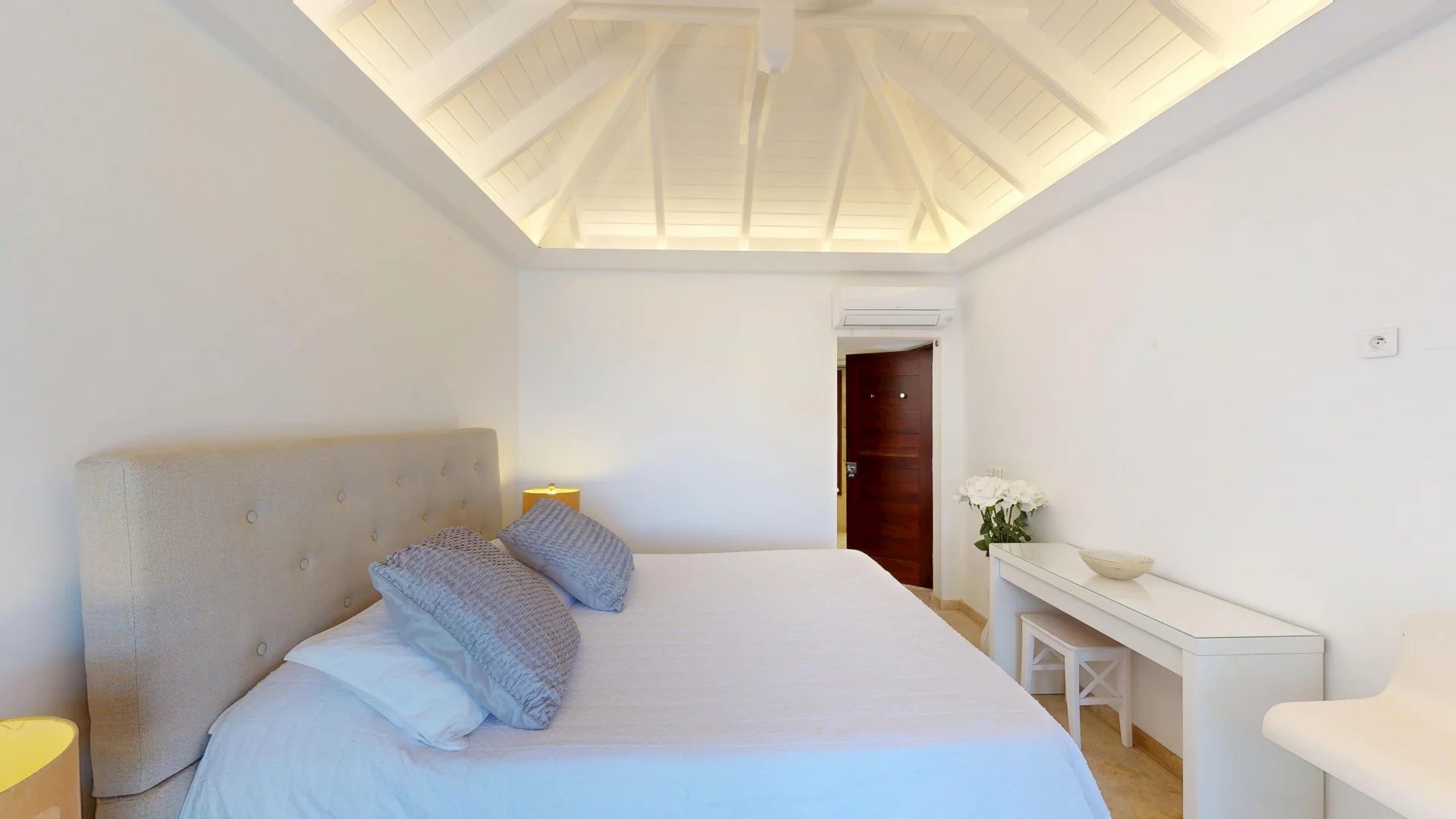 Bright minimalist bedroom with a vaulted white ceiling, beige upholstered bed, and gray pillows; door and desk with flowers against white walls.