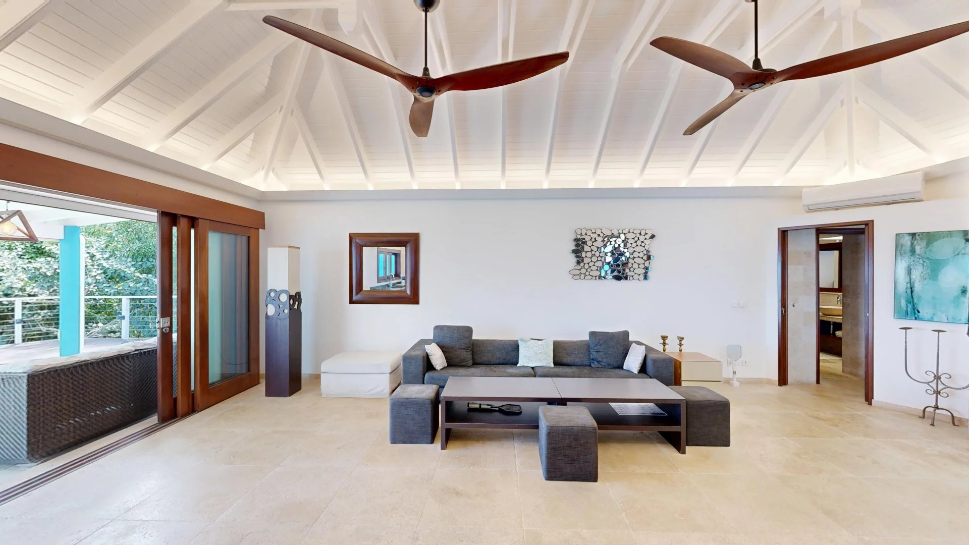 Bright living room with a white vaulted ceiling, two wooden ceiling fans, a gray sectional sofa, and sliding glass doors to a deck outdoor area.