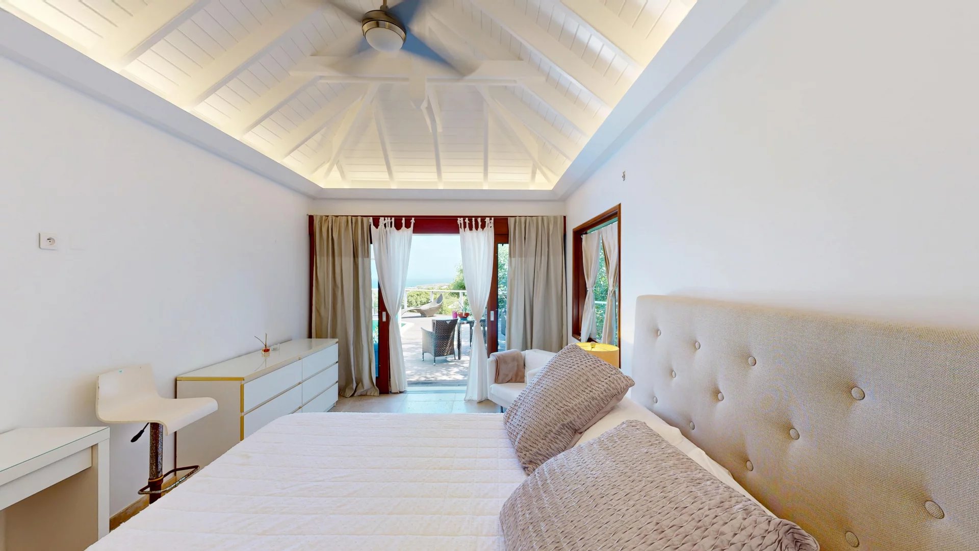 Bright bedroom with a white vaulted ceiling and a large tufted headboard, beige bedding, and balcony doors opening to an outdoor seating area.