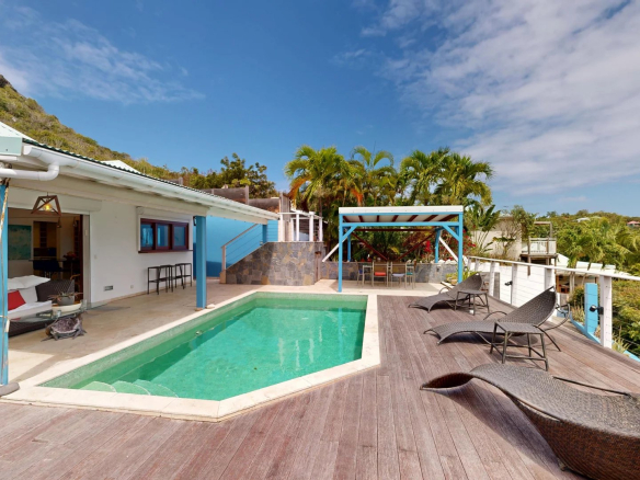 Poolside patio of a tropical villa turquoise pool wooden deck wicker loungers and palm trees under a blue sky hillside in background Propriétés clovis