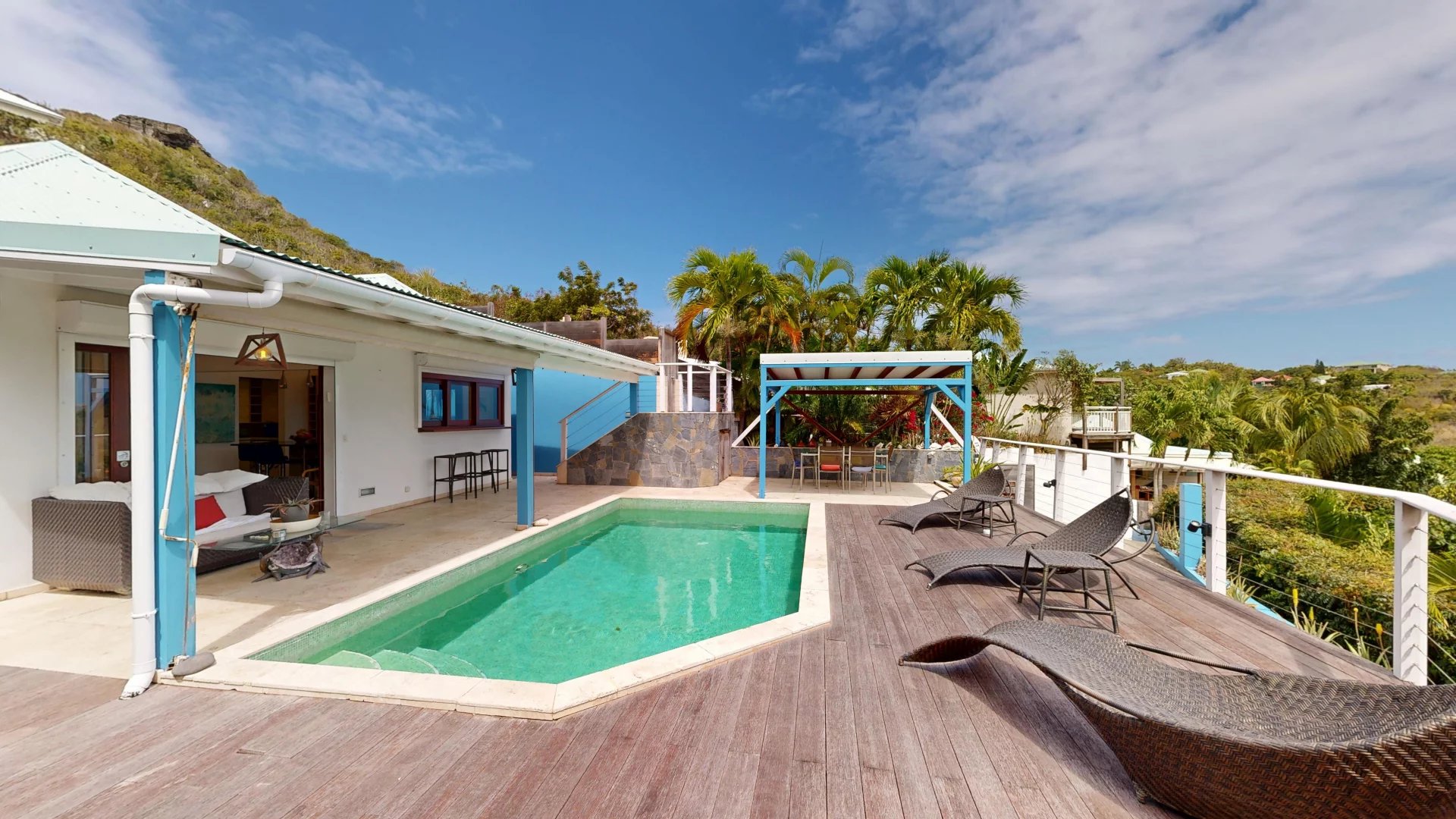 Poolside patio of a tropical villa turquoise pool wooden deck wicker loungers and palm trees under a blue sky hillside in background Propriétés clovis