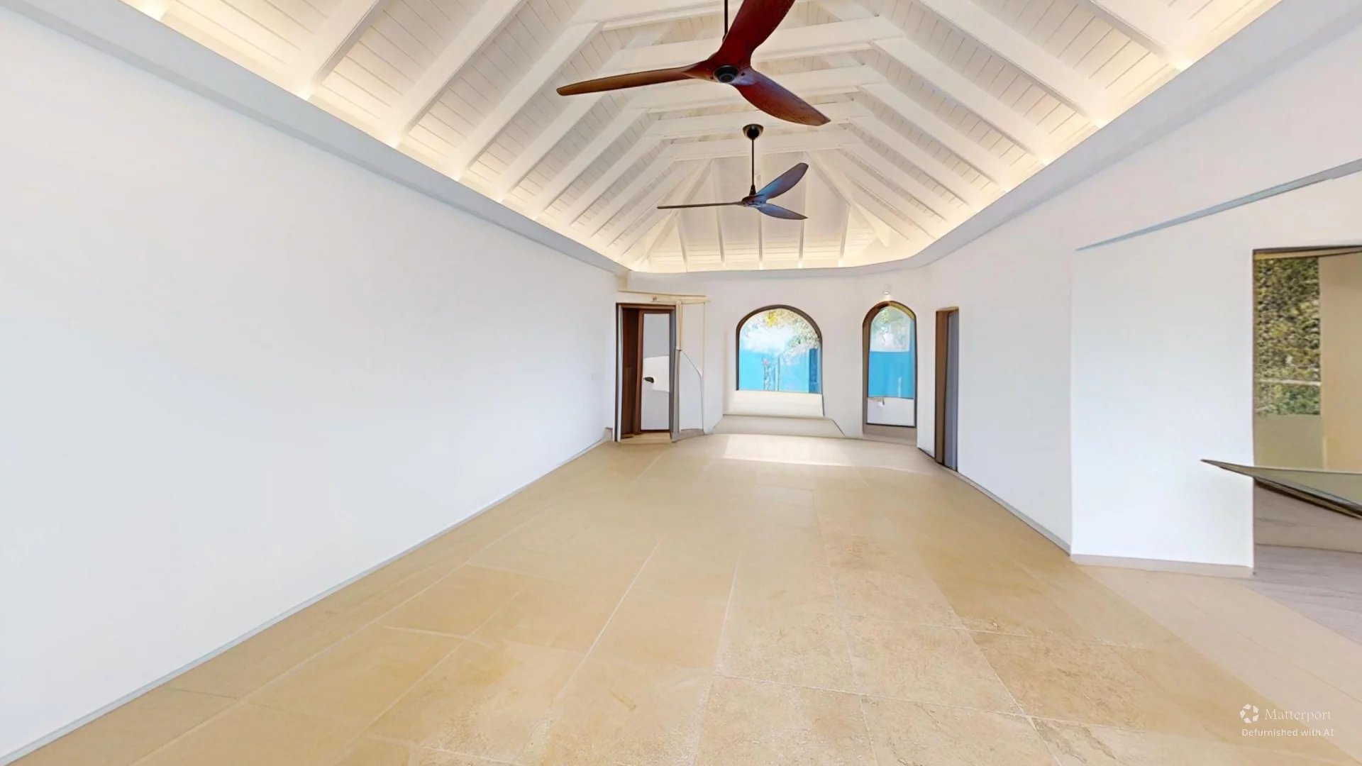Bright, empty room with white walls and a vaulted wooden ceiling, two ceiling fans, and arched doorways at the far end.