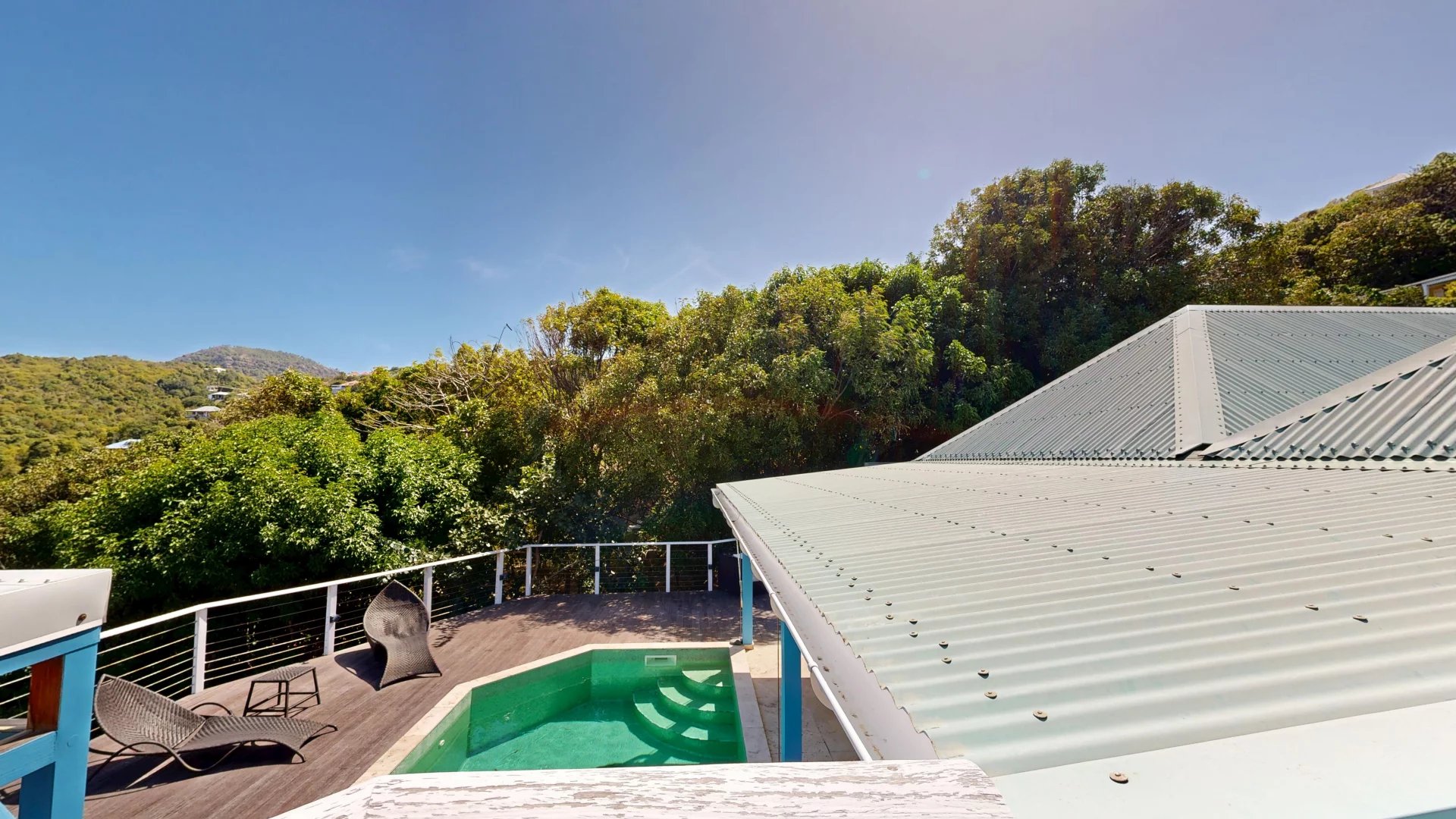 Rooftop deck with a small turquoise pool, two wicker lounge chairs, and white railing against a backdrop of trees and hills under a blue sky.