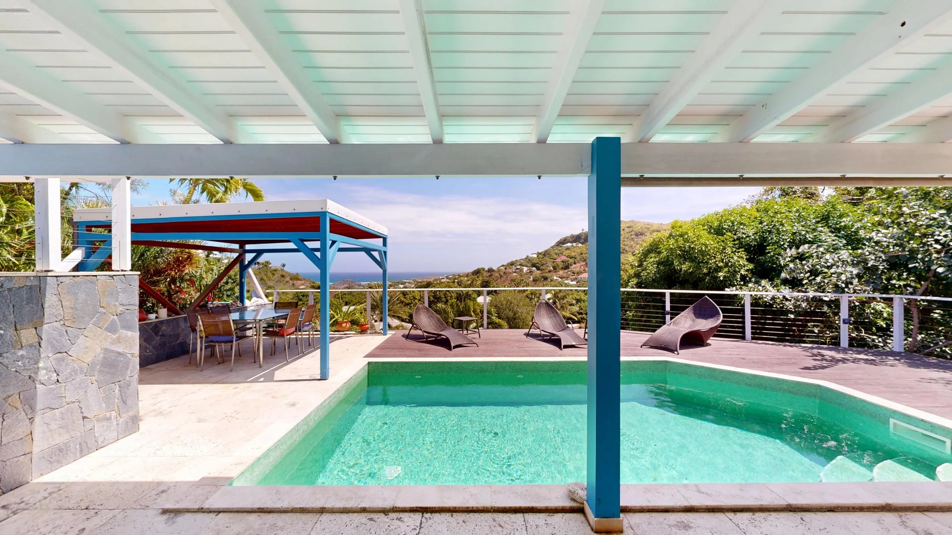 Outdoor pool area with a turquoise pool, stone wall, and a shaded dining area under a white-and-blue pergola. View of hills and sea beyond.
