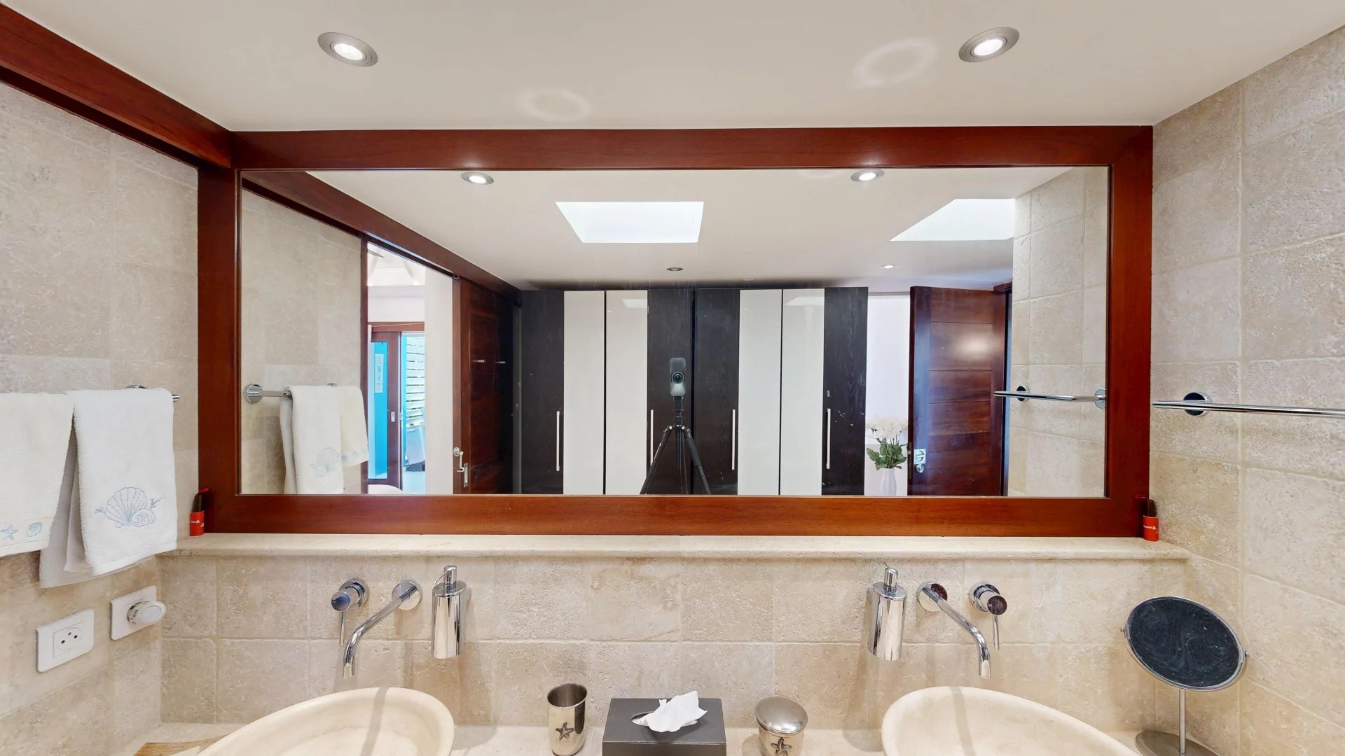 Modern beige-tiled bathroom with dual sinks and a large wooden-framed mirror; a camera on a tripod stands facing the mirror.