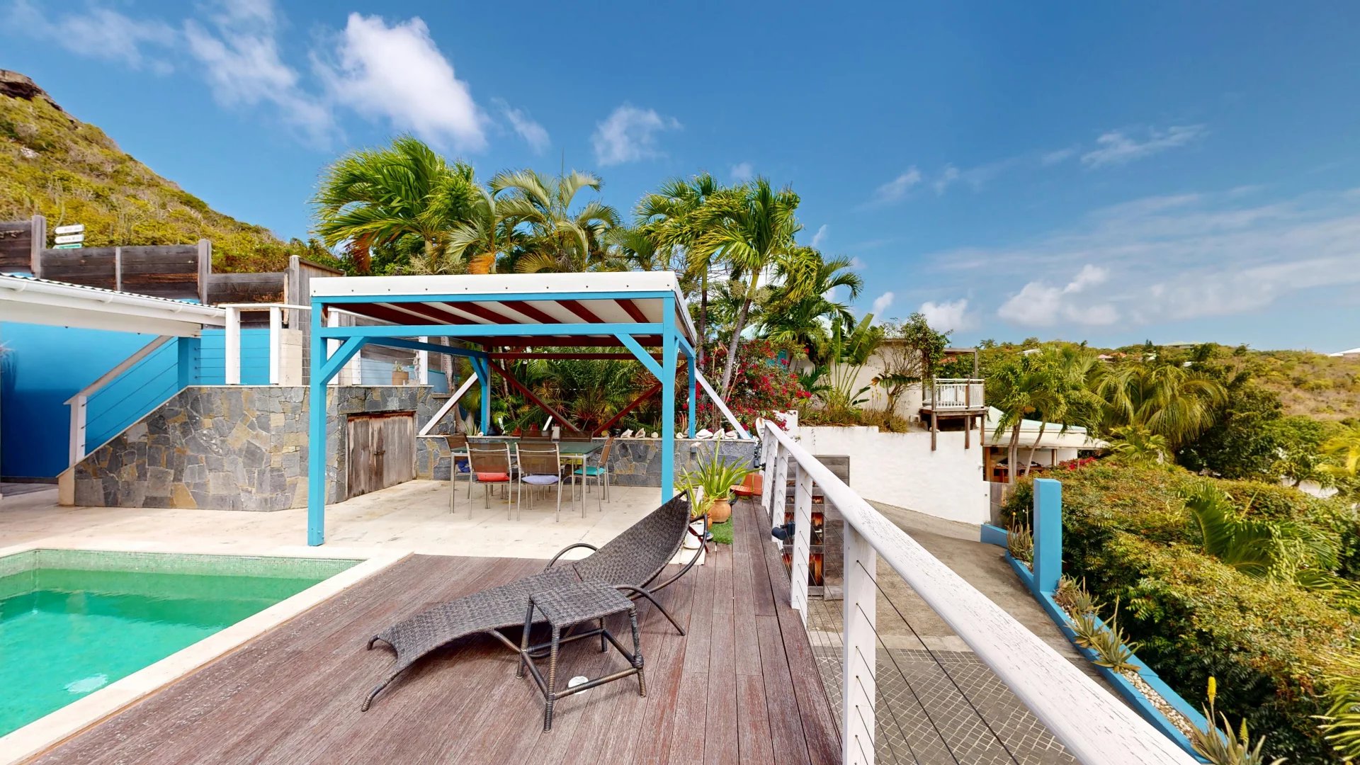 Poolside patio with a turquoise pool, wooden deck, lounge chair, and a blue-framed pergola among palm trees.