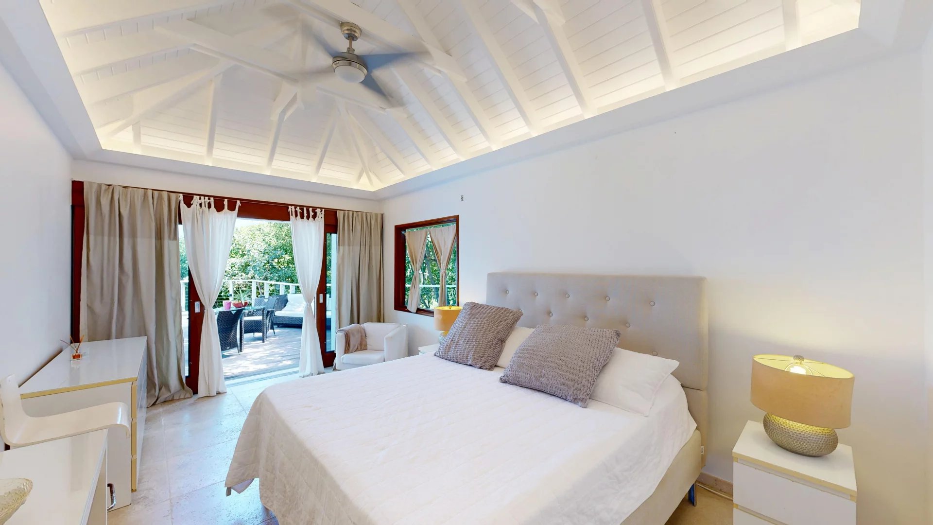 Bright bedroom with a large white bed and tufted headboard, white bedding, and matching nightstands; doors open to a sunlit balcony. Next to a seating area by the window.