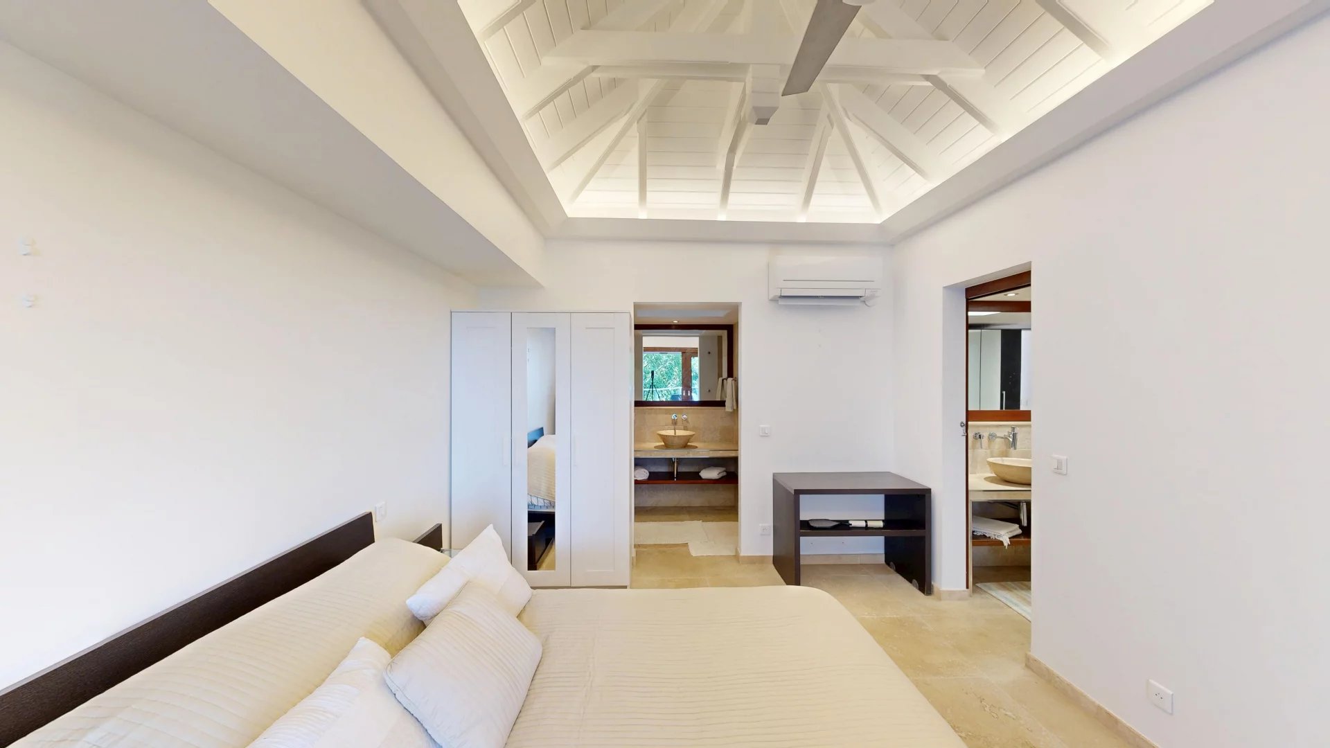 Bright white bedroom with a vaulted ceiling and exposed beams, a large bed with beige bedding, a white wardrobe, and an en-suite doorway visible ahead.