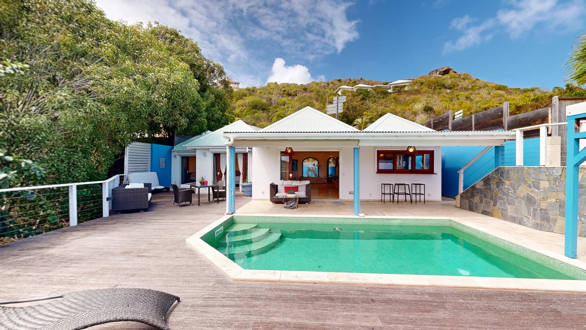 Villa with a turquoise pool, white patio, and outdoor seating on a wooden deck under a blue sky.