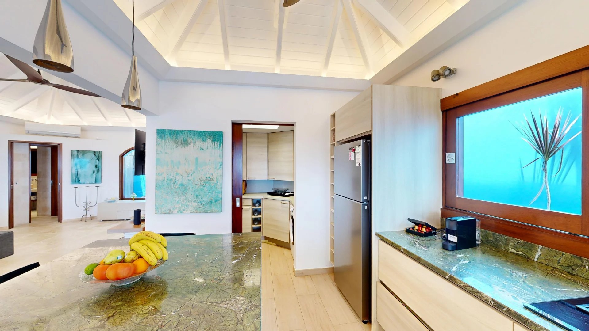 Bright open-concept kitchen with a green marble island, fruit bowl, and stainless steel refrigerator under a vaulted white ceiling.