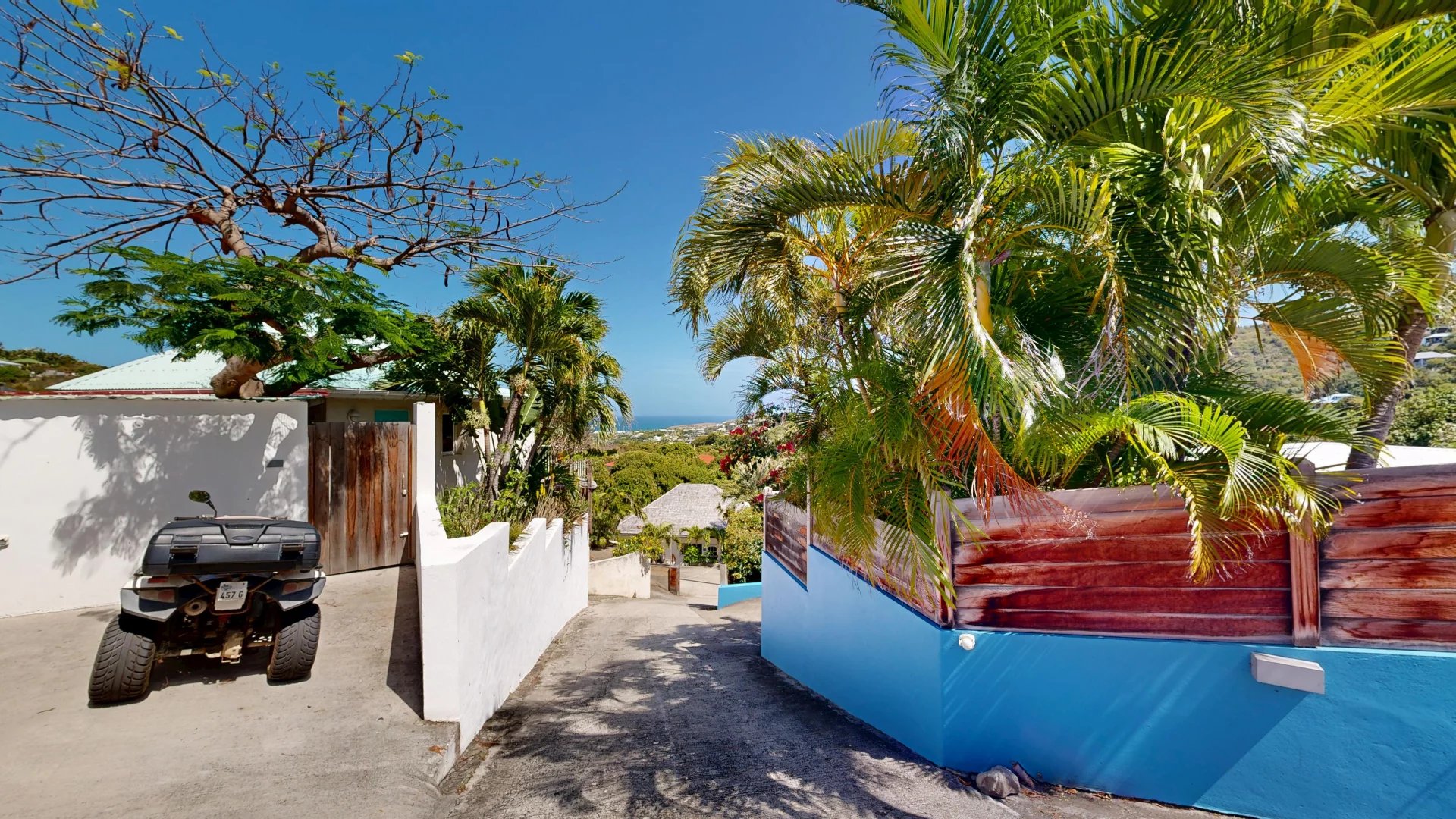 Quad bike parked along a sunlit tropical street with white walls, palm trees, and a view of the sea in the distance.