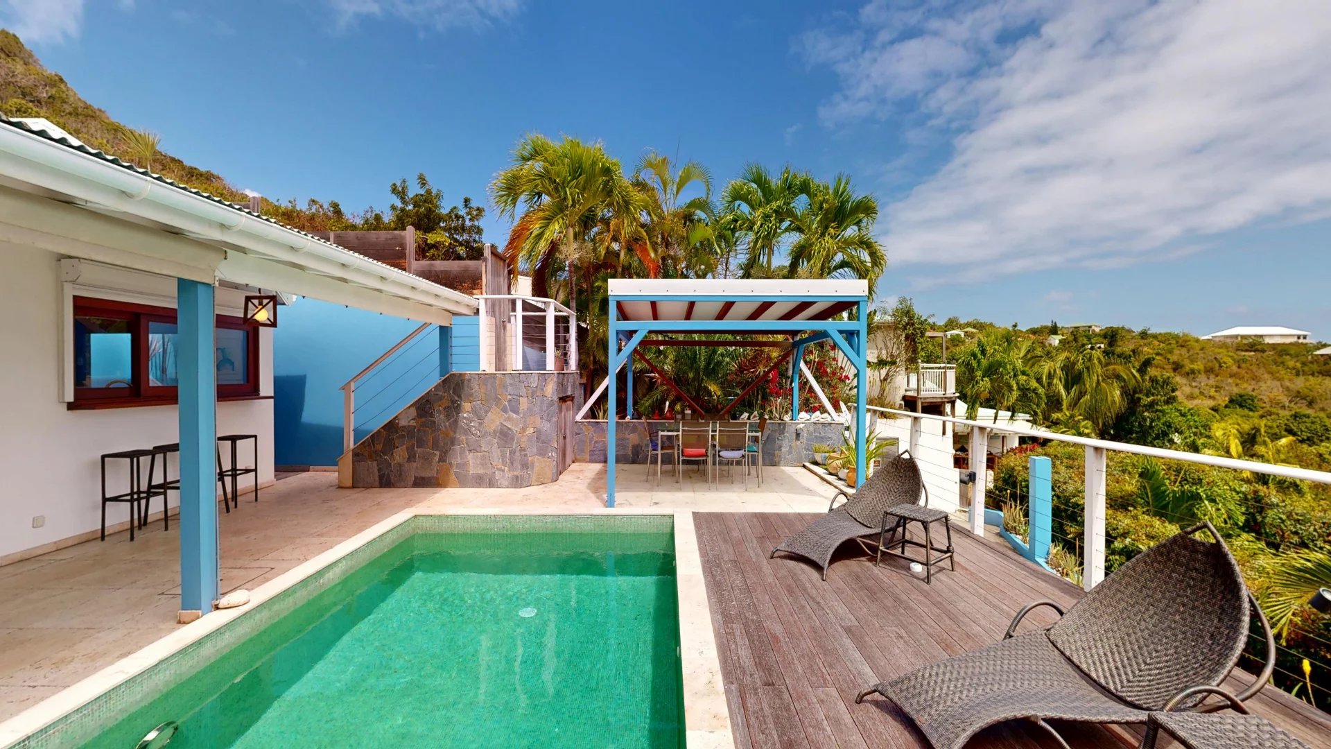 Outdoor pool area with turquoise water, wooden deck, and wicker lounge chairs under a bright blue sky.