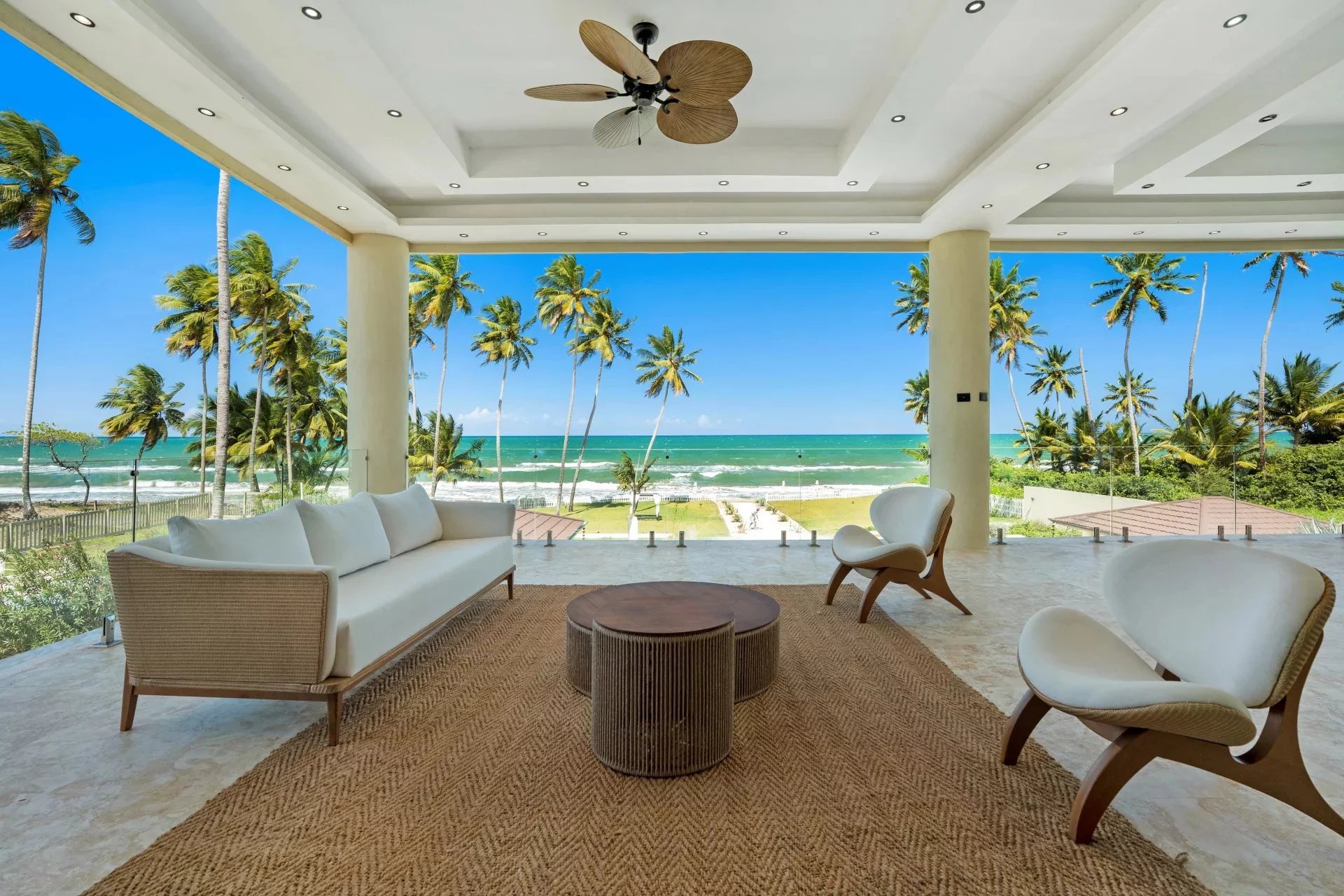 Open-air living area with beige wicker sofa and chairs, overlooking a tropical beach and palm trees.