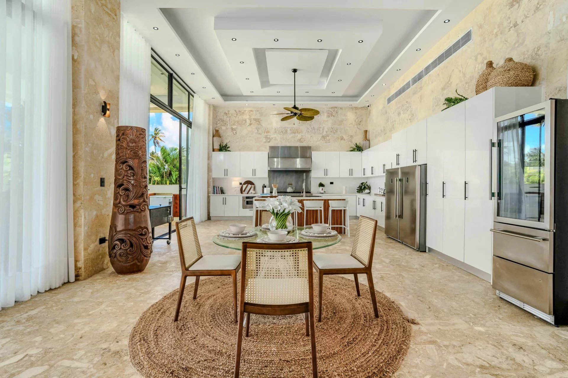 Open-plan kitchen and dining area with white cabinets, stainless steel appliances, and a round glass dining table on a woven rug near large windows.