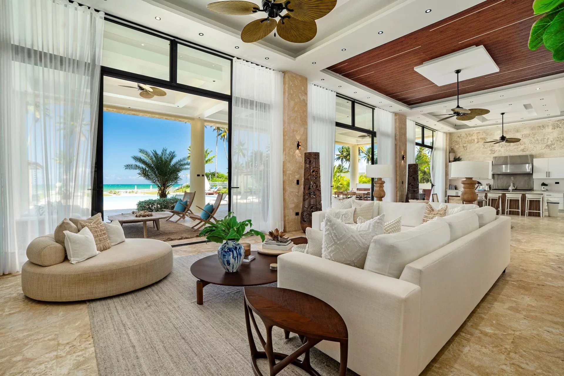 Luxurious open-plan living room with white sofas, beige chaise, and a view of the beach through glass doors.