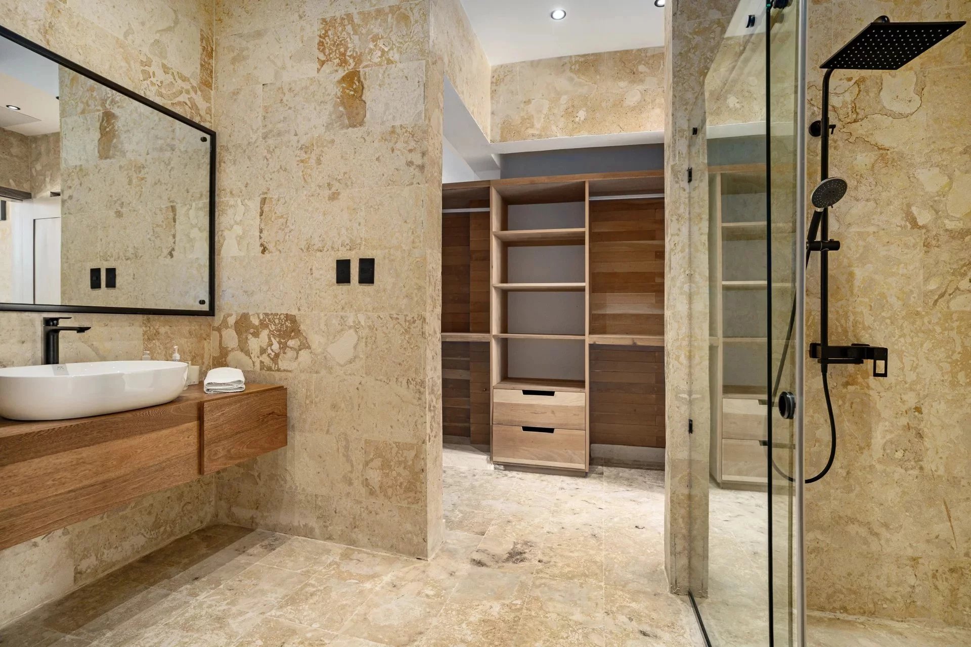 Modern bathroom with beige stone walls, a wall-mounted wooden vanity, and a white vessel sink beneath a large mirror.