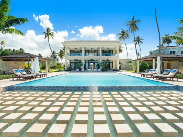 Luxurious resort pool scene with a square stepping stone path across shallow water toward a modern two story building and palm trees under a blue sky Propriétés clovis