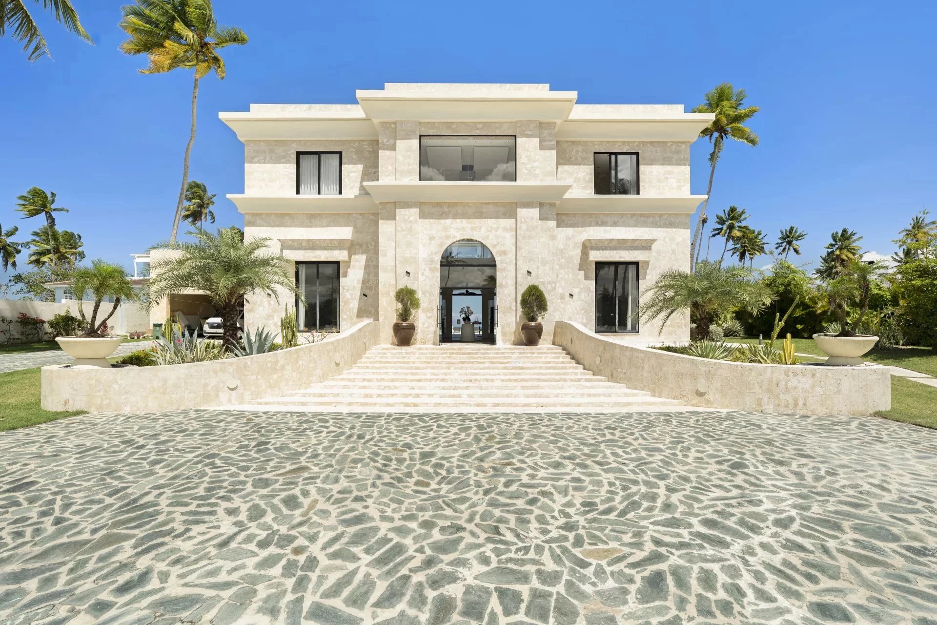 Luxurious light stone villa with a central arched doorway, wide stone steps, and palm-lined grounds.