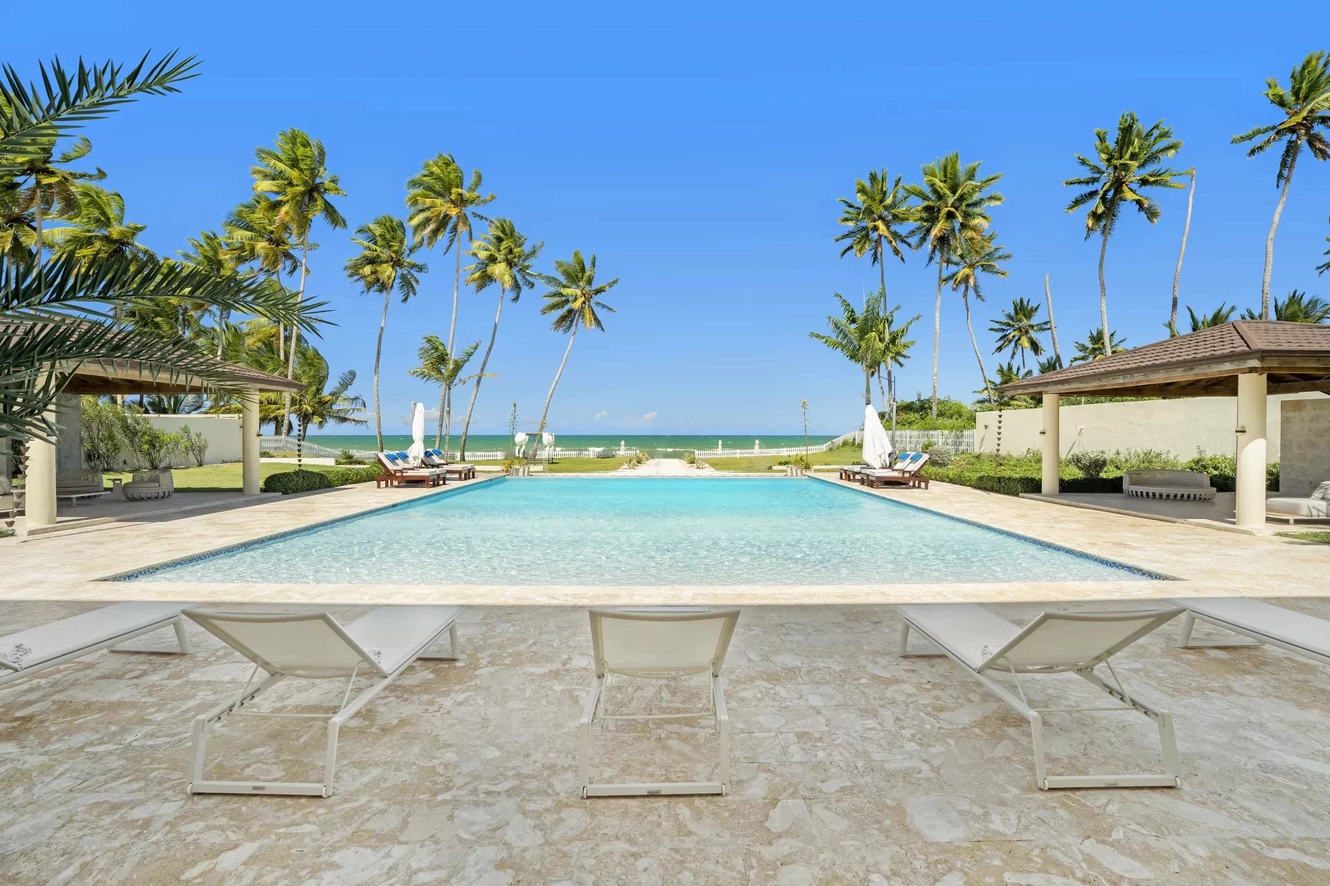 Resort pool surrounded by palm trees with lounge chairs and a view of the ocean on a sunny day