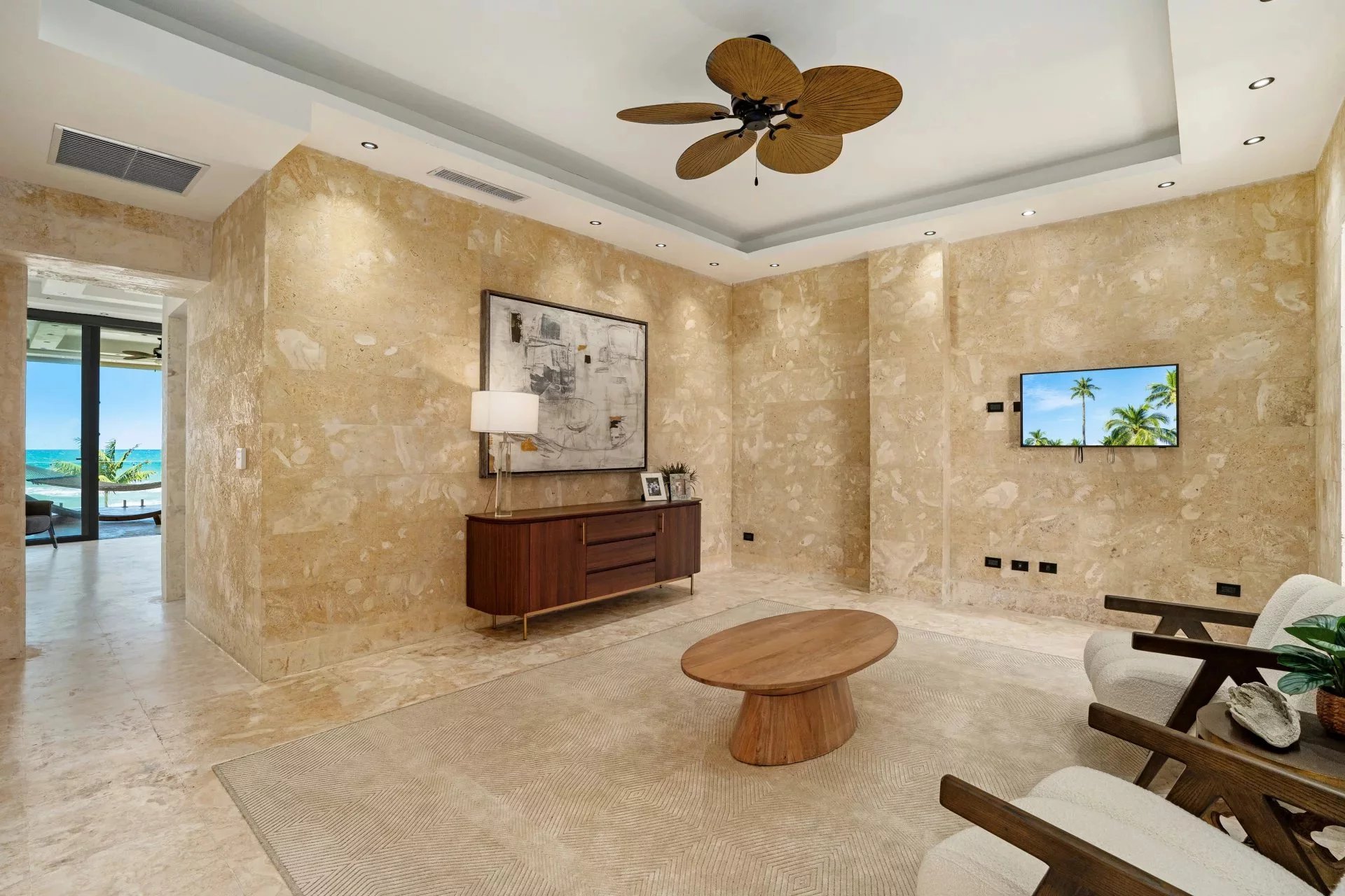 Luxurious beige marble living room with a wooden console, abstract wall art, and a ceiling fan; view to a sunny beach through an open doorway on the left
