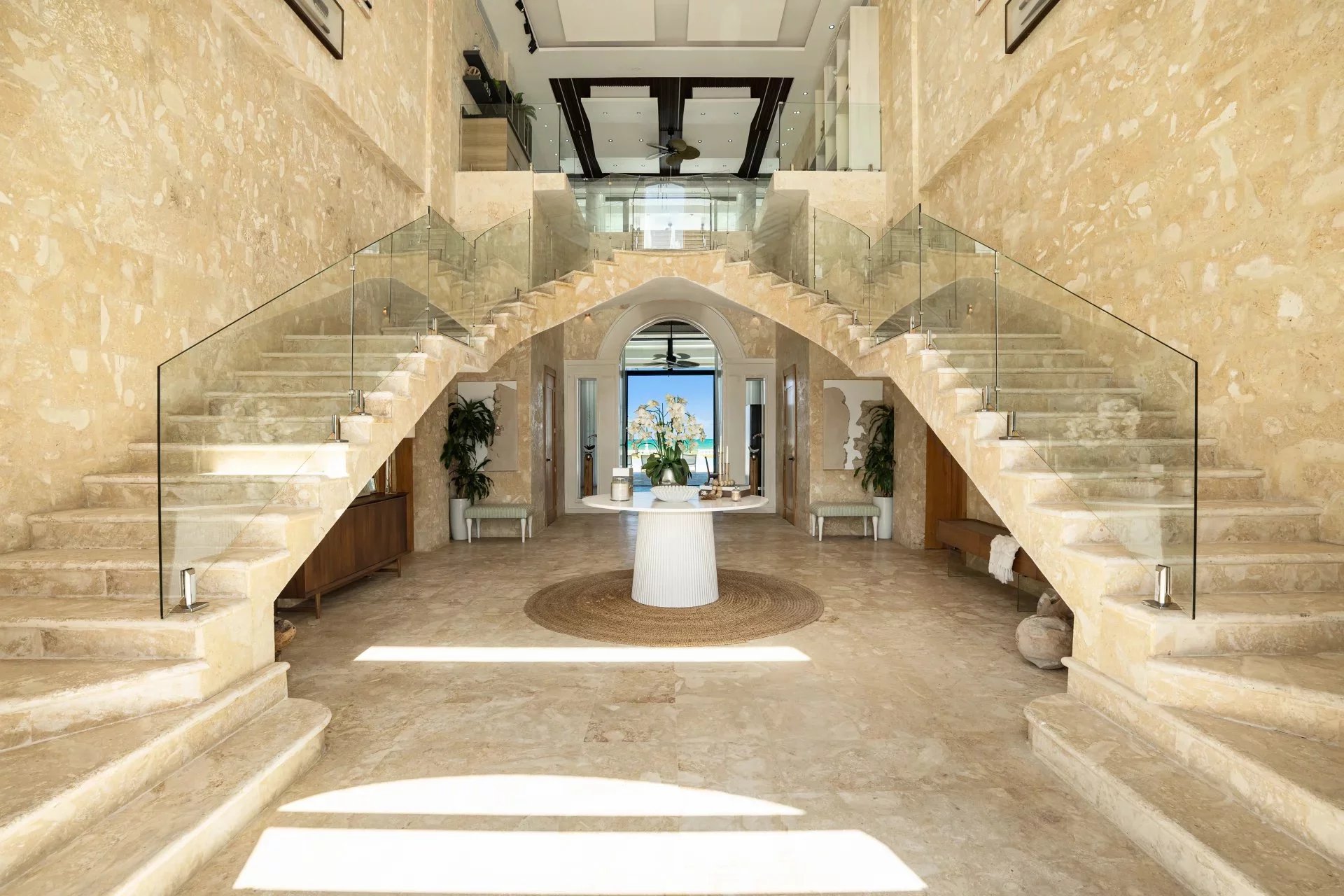 Grand marble foyer with double stone staircases and glass railings, central round table, and ocean view through an arched doorway.