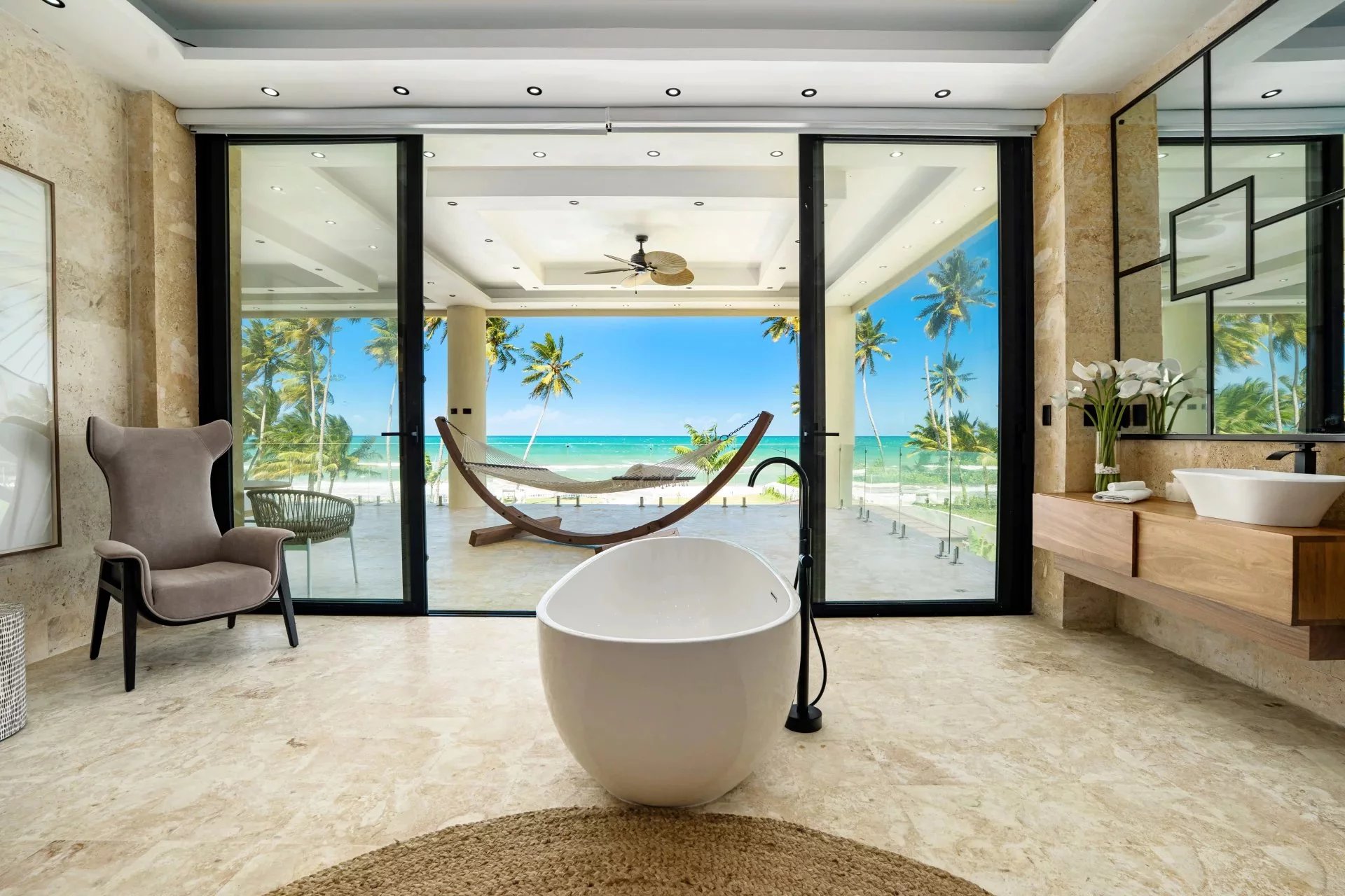 Luxurious bathroom with a freestanding white tub facing a tropical beach view through glass doors and a hammock on the patio beyond.