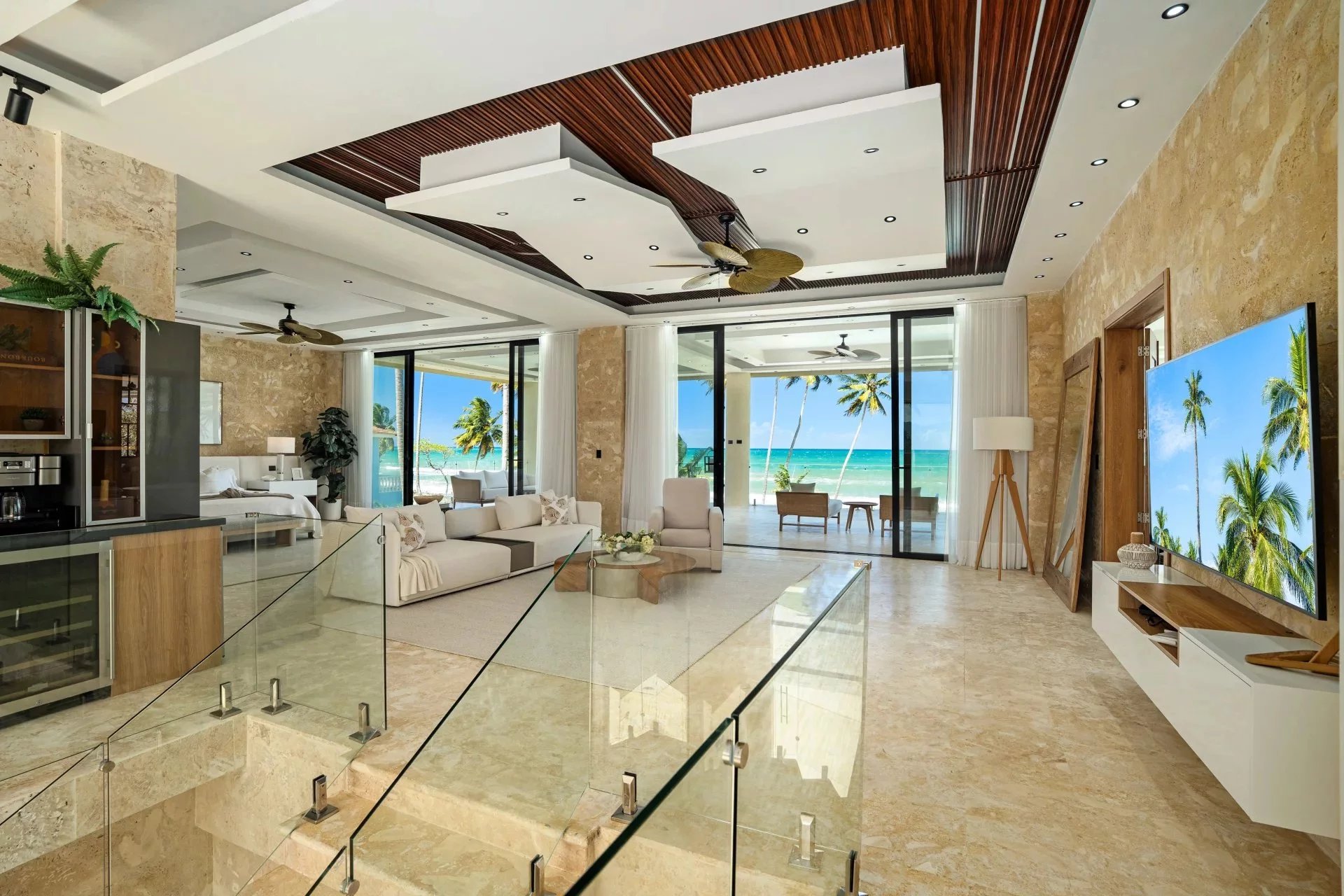 Luxurious open-concept living room with beige marble floors, glass railing, and a view of palm trees and the ocean through sliding doors
