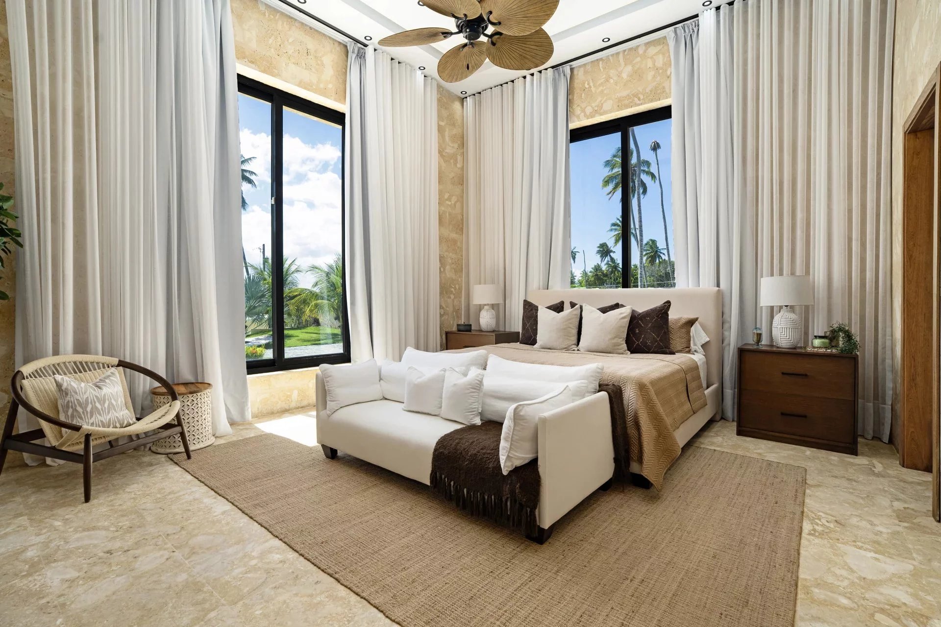 Luxurious bedroom with a white bed, beige bedding, and multiple pillows, flanked by dark wood nightstands and large windows revealing palm trees outside