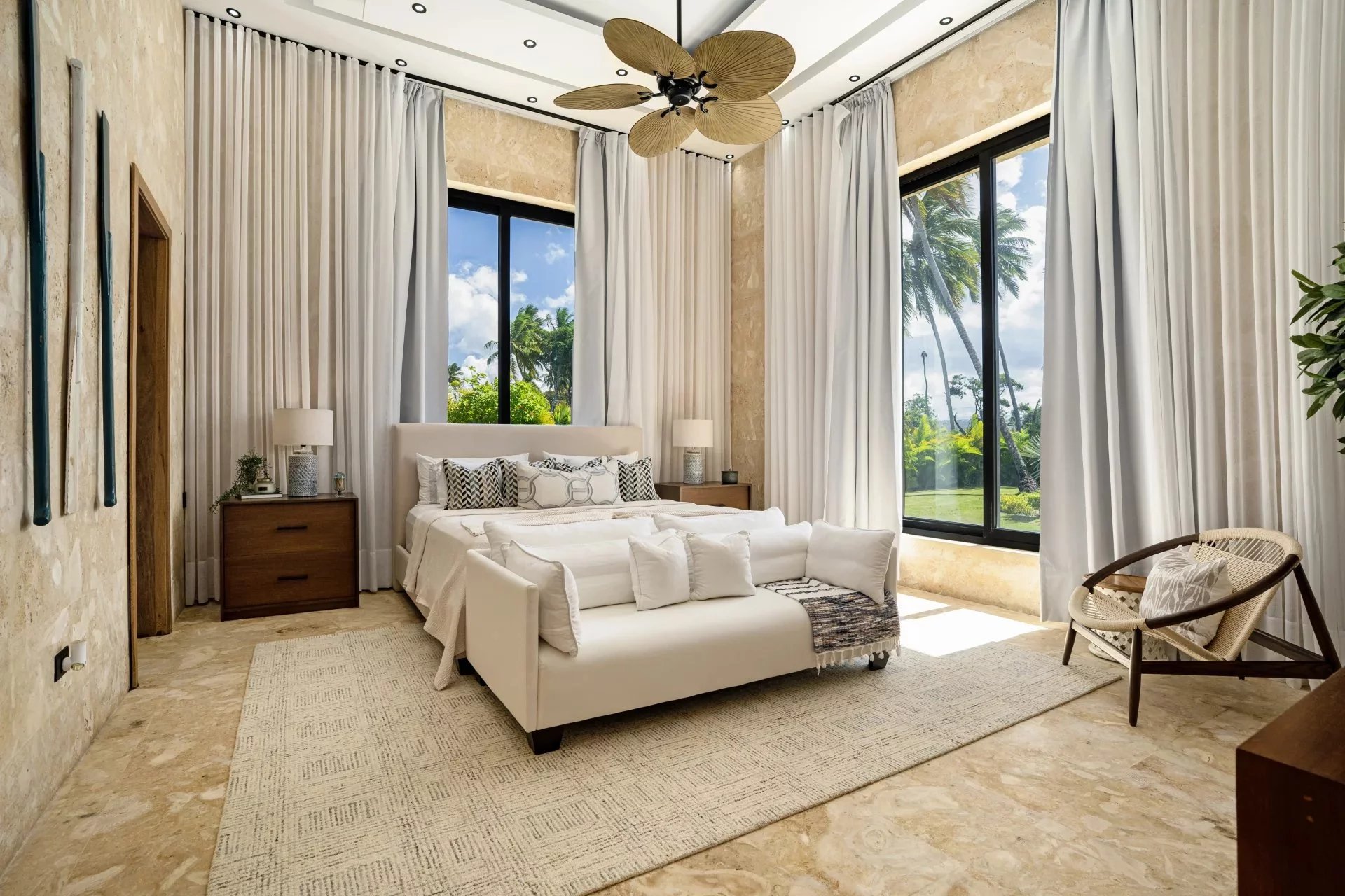 Luxurious beige-toned bedroom with a white bed, sofa at the foot, and large windows overlooking palm trees.