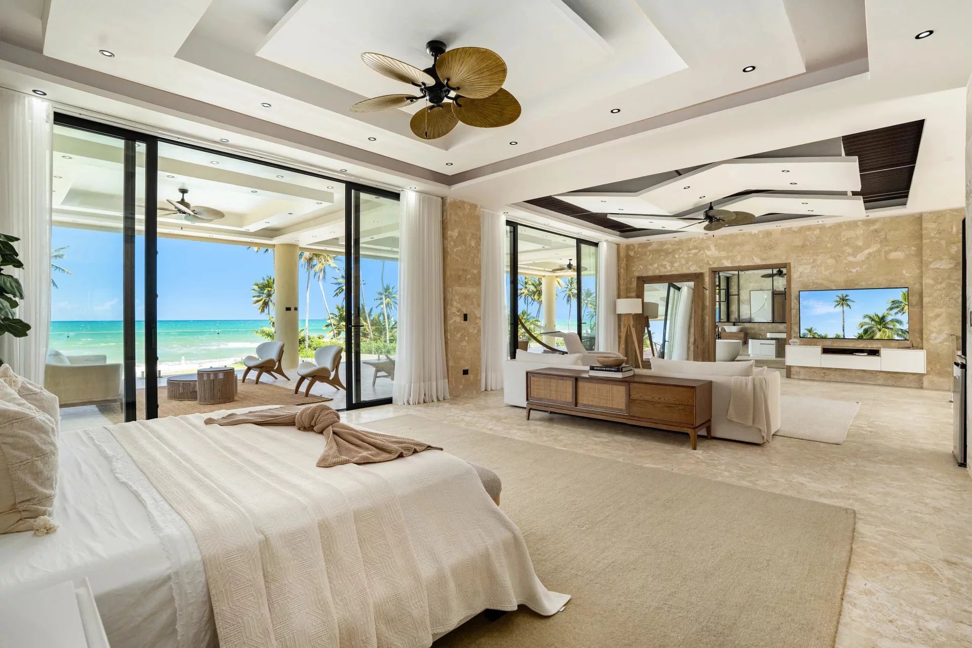 Luxurious beachfront bedroom with sliding glass doors open to an ocean view and palm trees on a sunny balcony.