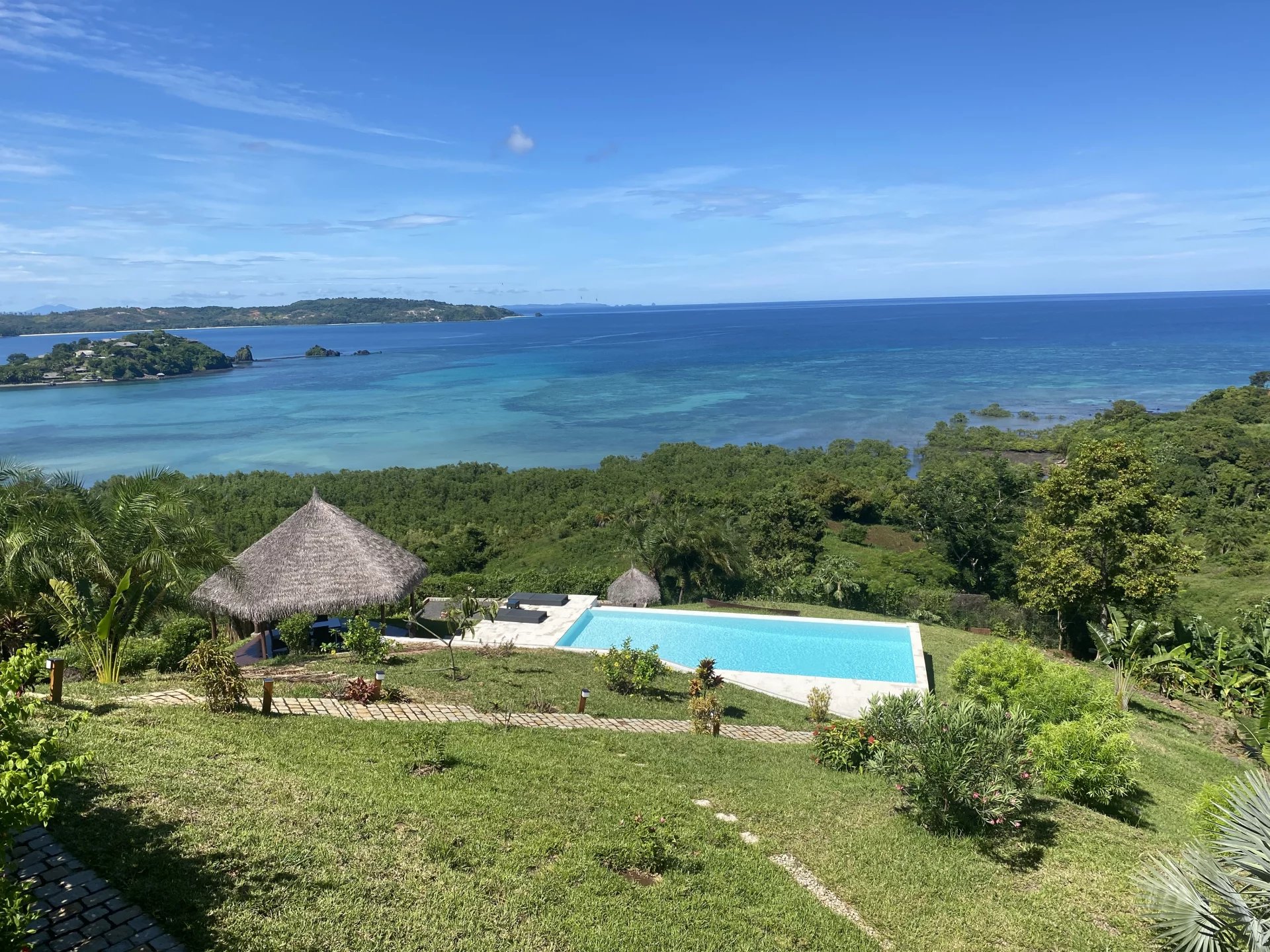 Resort terrace with a rectangular pool overlooking turquoise sea and lush greenery under a clear blue sky