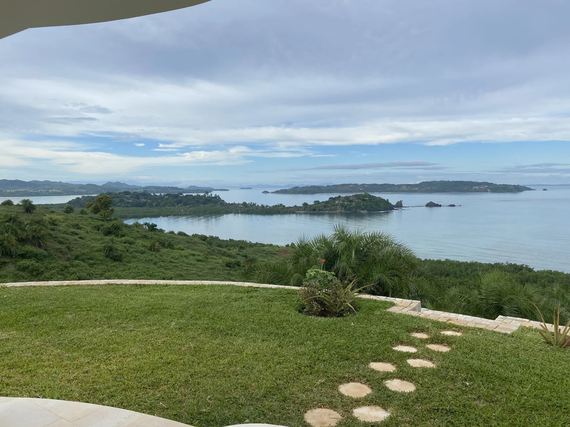 Coastal landscape with calm blue water, small islands, green hills, and a grassy foreground with stepping stones beneath a cloudy sky.