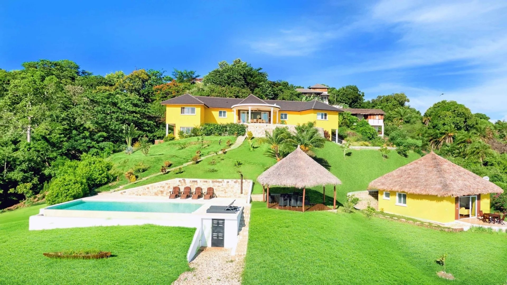 Yellow two-story villa on a grassy hill with a pool, lounge chairs, and a thatched gazebo; tropical landscaping and clear blue sky.