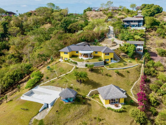 Aerial view of a yellow two story villa with a curved stone terrace and shaded patio on a hillside surrounded by greenery and other buildings Propriétés clovis