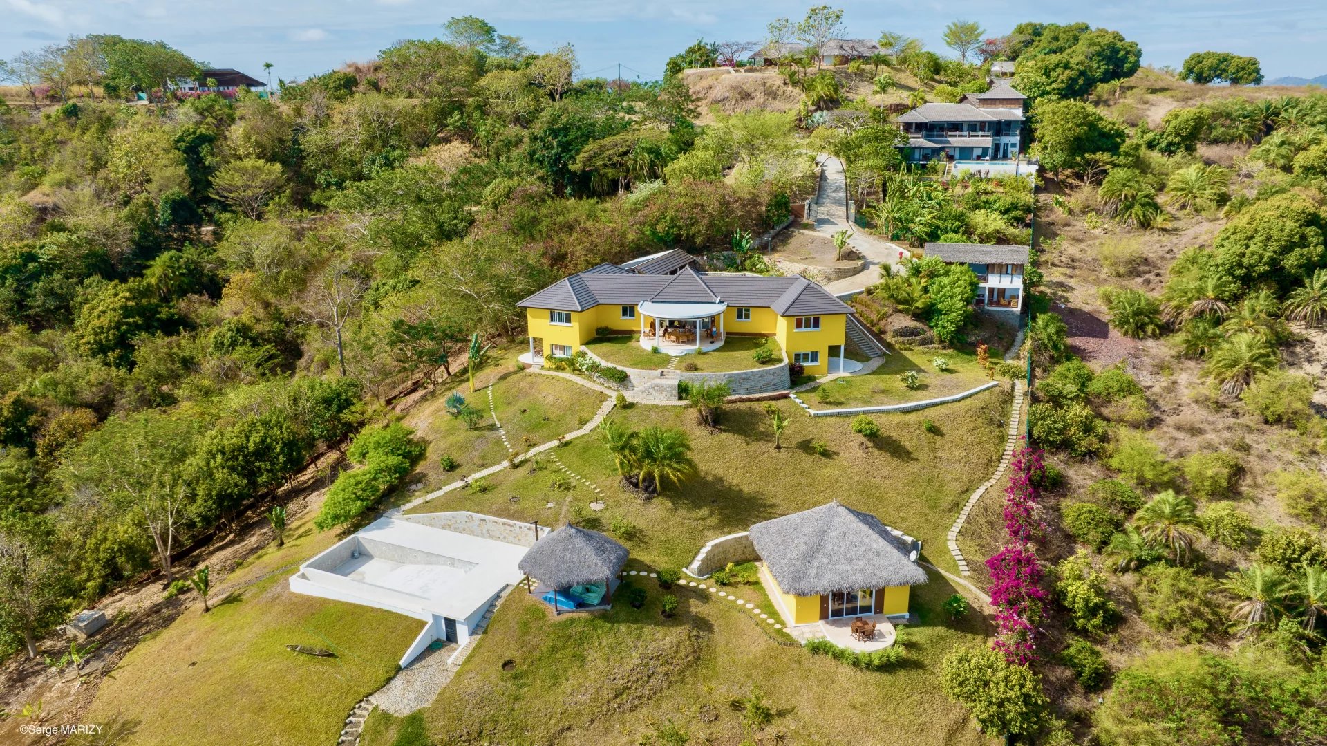 Aerial view of a yellow two story villa with a curved stone terrace and shaded patio on a hillside surrounded by greenery and other buildings Propriétés clovis
