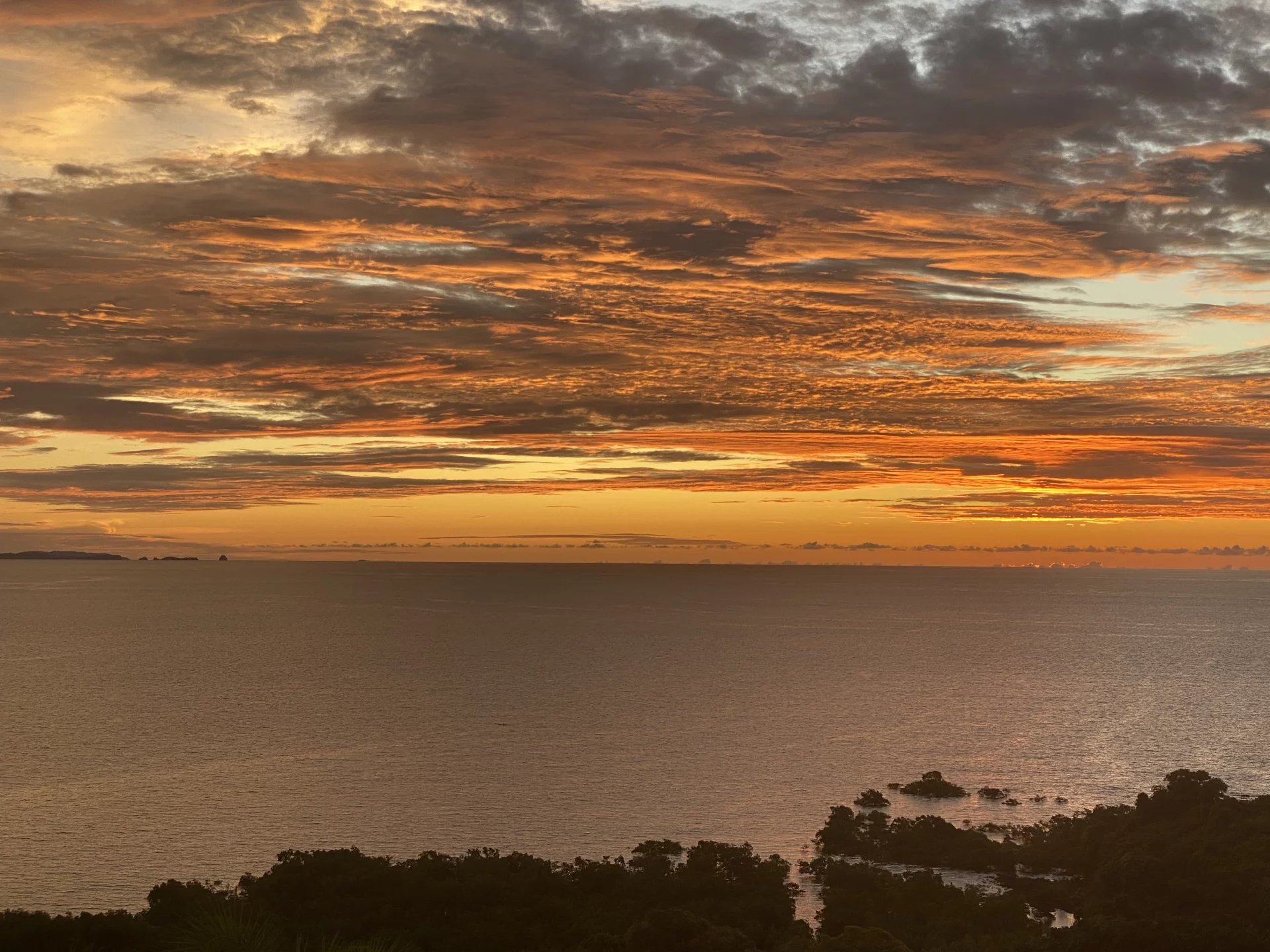 Sunset over a calm sea with orange clouds and dark coastal silhouettes in the foreground.",