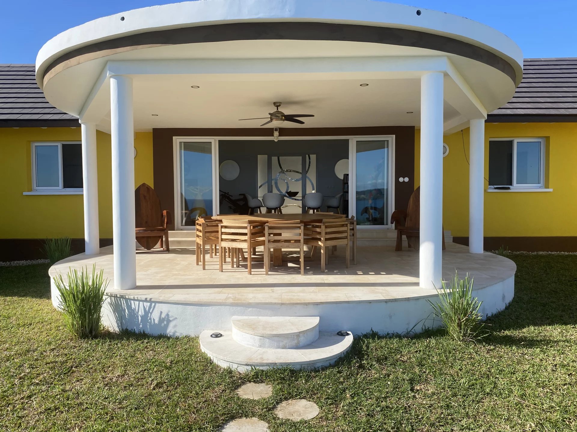Front porch of a bright yellow house with a curved white roof, white columns, and a wooden outdoor dining table and chairs on a tiled patio.