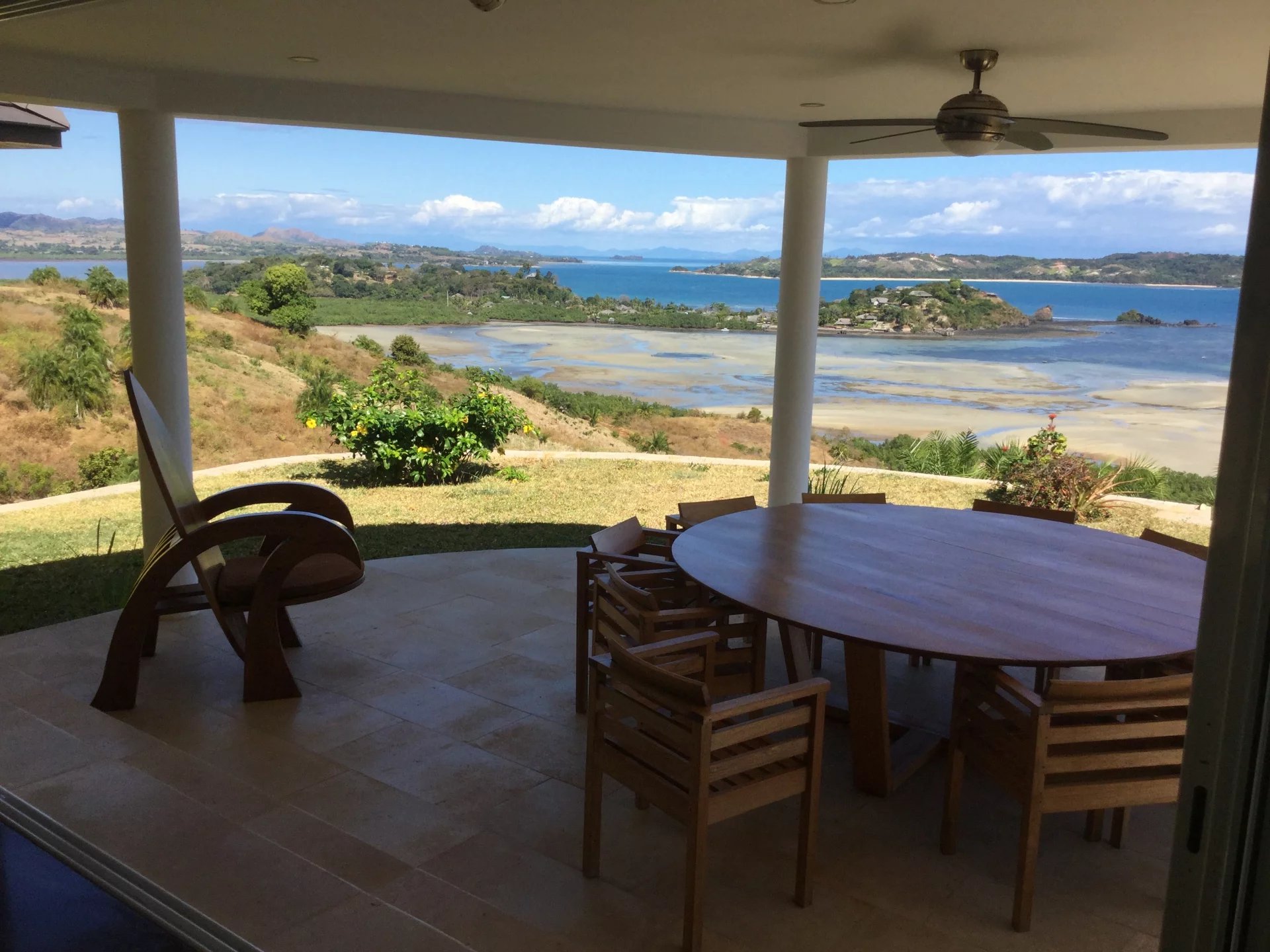 Covered patio with a round wooden table and multiple chairs, overlooking a coastal bay and small island in the distance under a blue sky.