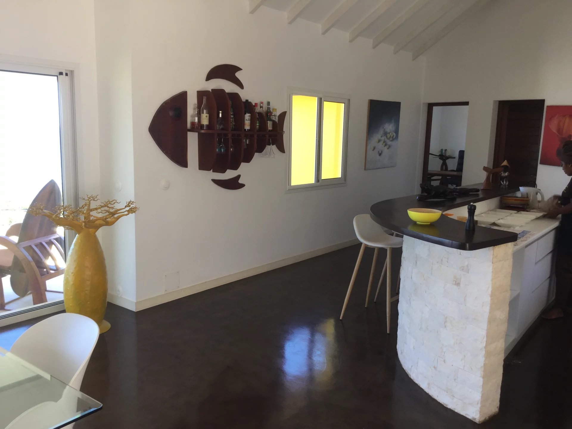 Open-plan kitchen with a curved stone bar, white walls, and a person working at the counter; decorative bottle rack on the wall.
