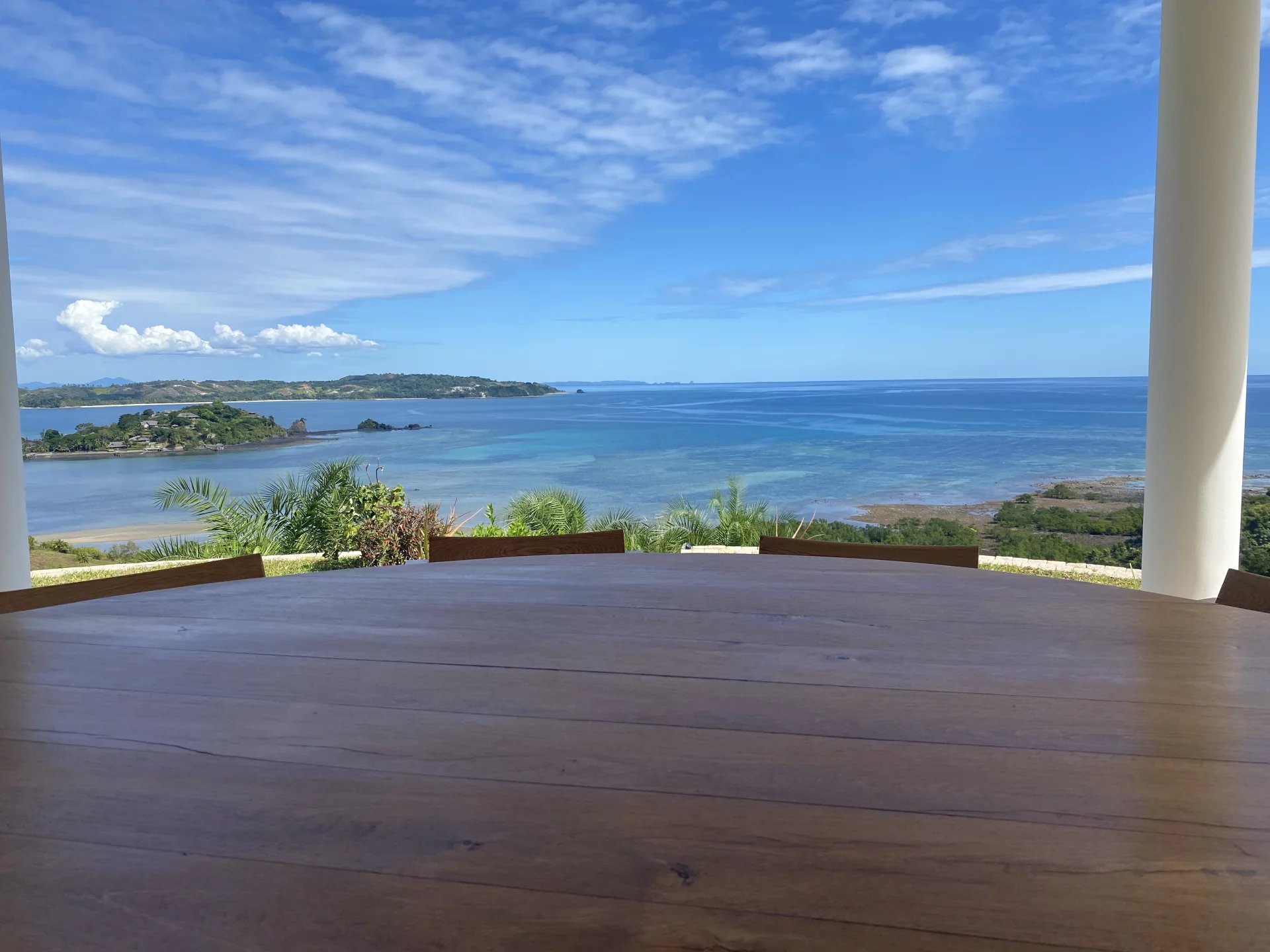 Outdoor dining table on a covered veranda overlooks a turquoise sea and distant islands under a clear blue sky, with palm plants nearby.