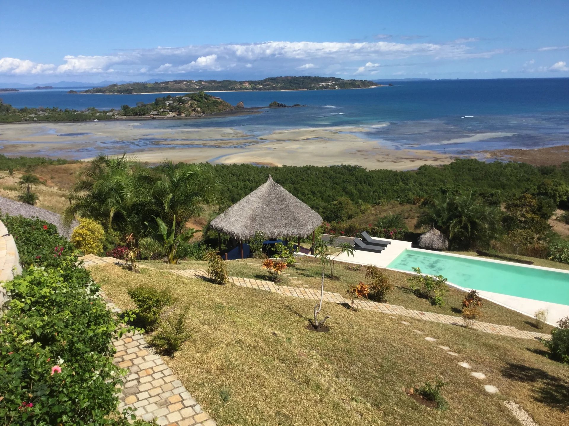 Resort view: thatched cabana, loungers, and a turquoise pool on a grassy hillside overlooking a blue ocean and distant islands.