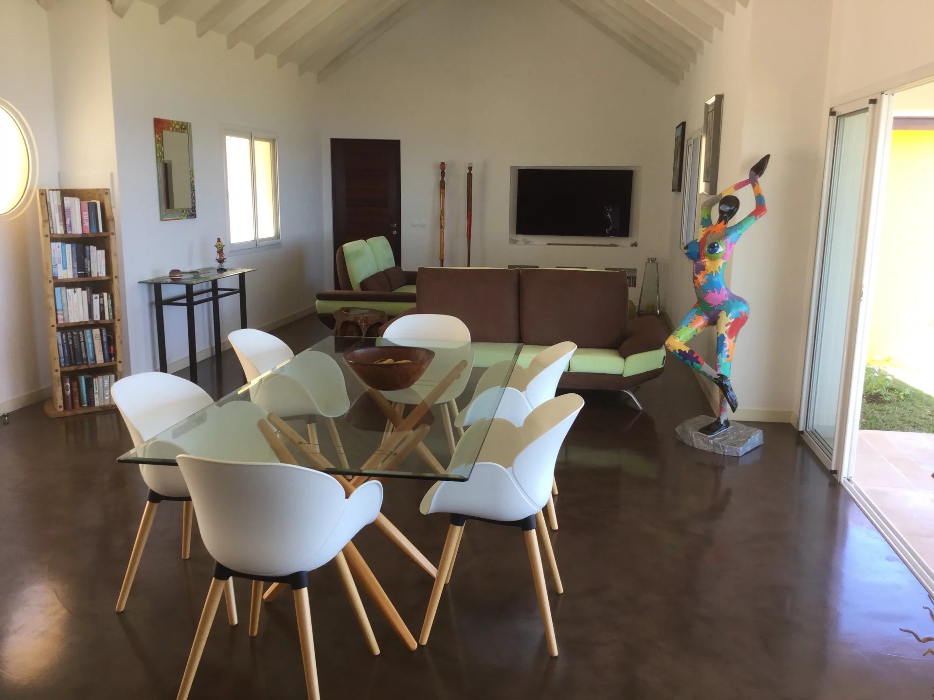 Open-plan living and dining area with a glass dining table surrounded by white molded chairs; a colorful abstract sculpture stands by the sliding glass door.