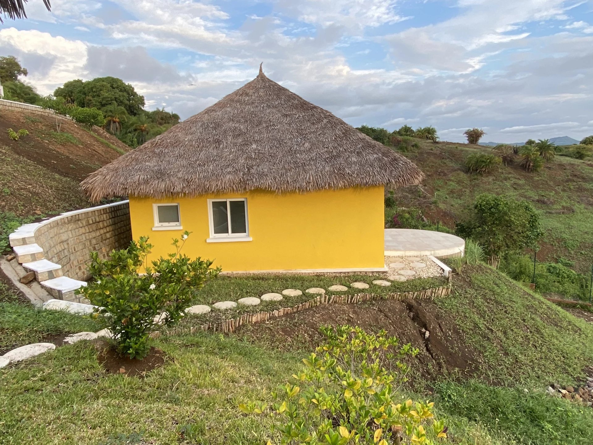 Yellow cottage with a thatched roof on a grassy hillside, bordered by a curved stone wall and circular patio edge.