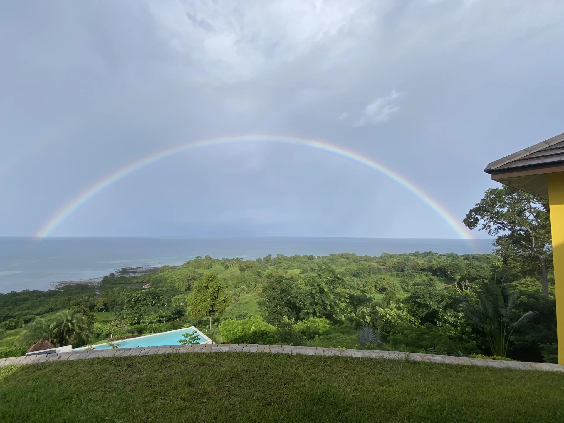 Double rainbow arcing over a green coastal landscape with the ocean in the distance, a pool in the foreground, and a yellow building corner on the right.