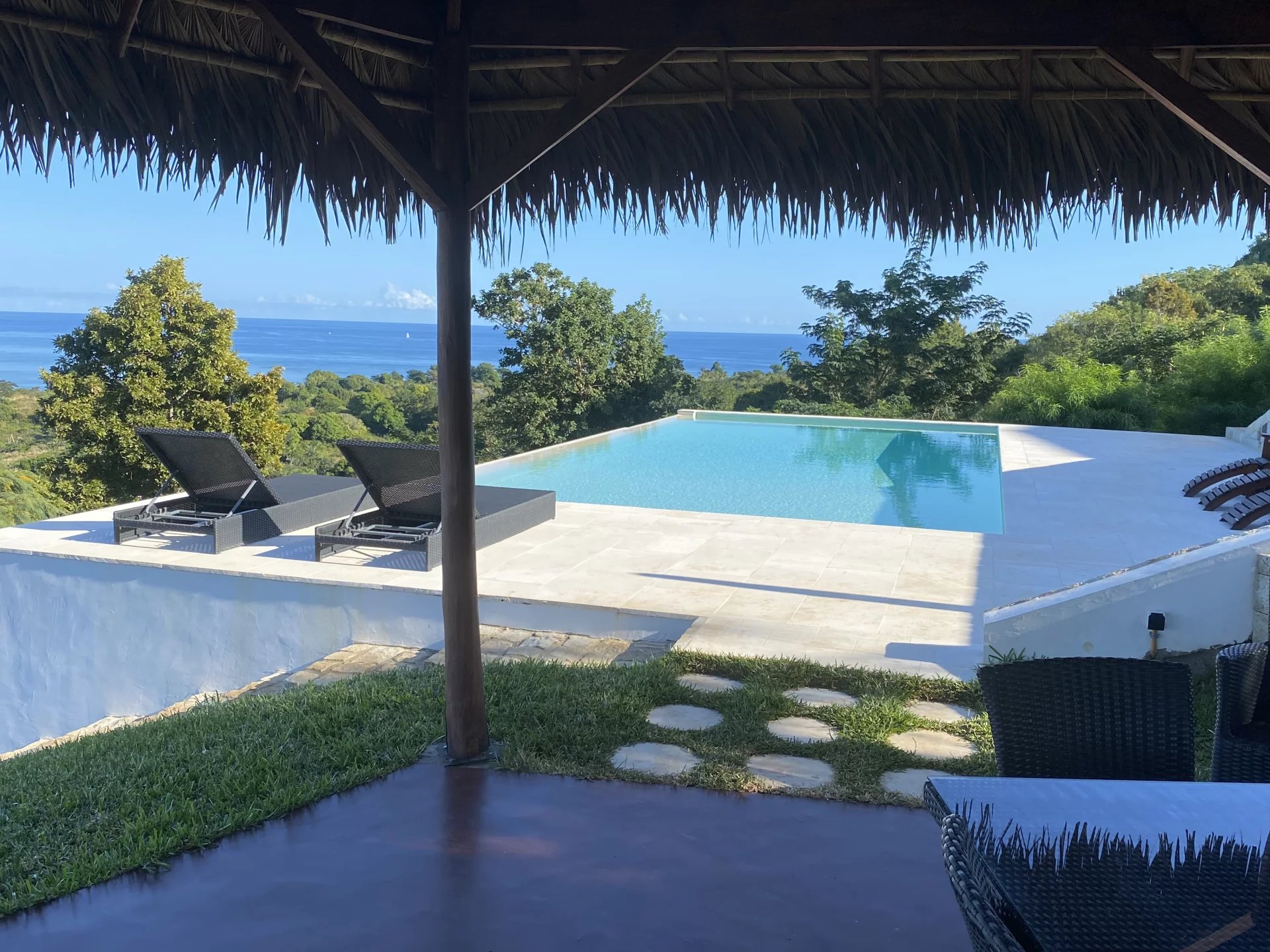 Outdoor infinity pool on a tiled deck with two black wicker lounge chairs, framed by a thatched roof and lush trees, overlooking the ocean.